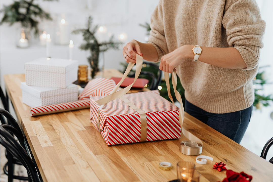Person wrapping a striped gift box with ribbon on a wooden table, surrounded by holiday decorations and wrapped presents.