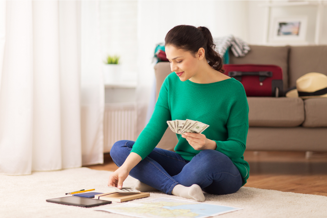 Woman sitting on the floor counting cash and organizing money next to a tablet, notebook, and map in a living room.