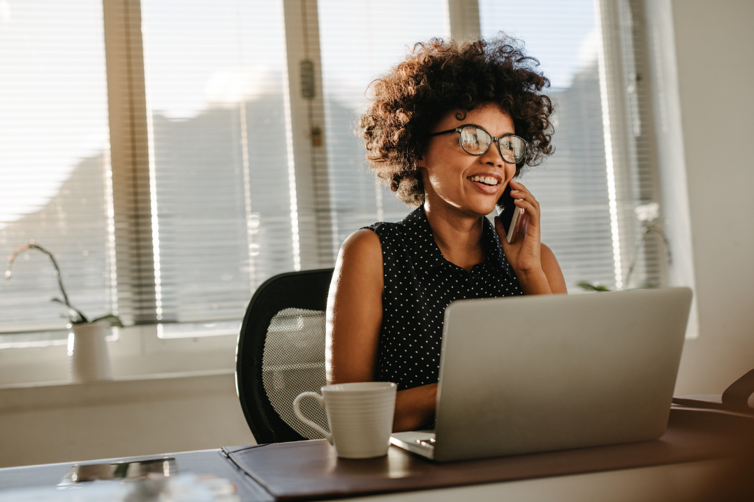 Woman working at a desk, smiling while talking on the phone and using a laptop in a bright office.