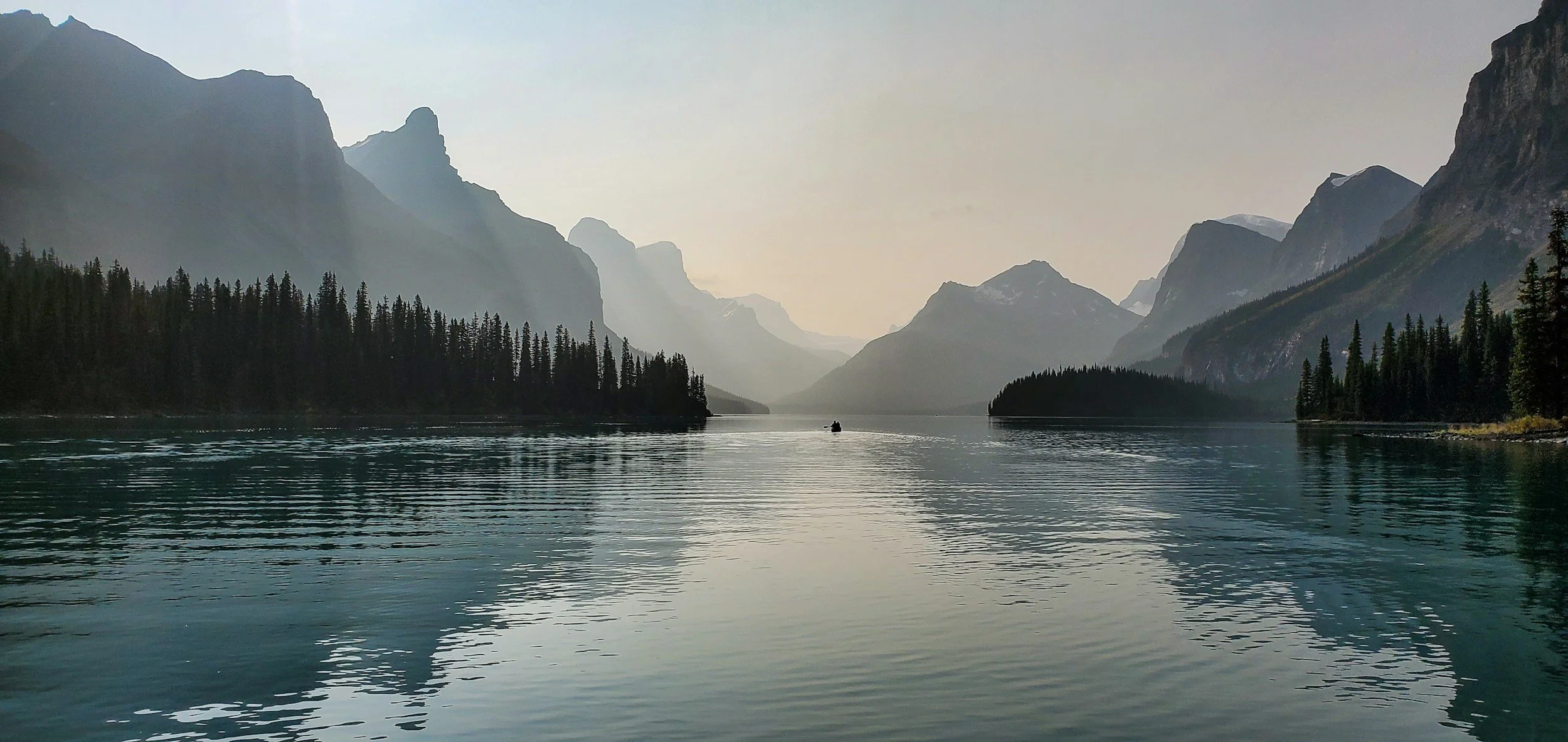 A serene body of water surrounded by tall pine trees and towering mountains in the distance, with a small boat floating on the water.