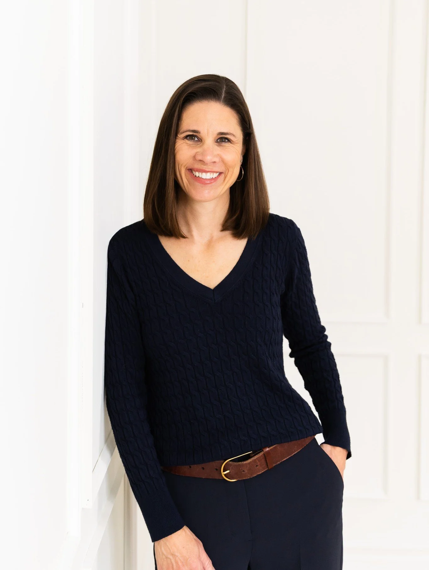 Tanya Melykuty, therapist, smiling, wearing a navy blue knitted sweater and black pants, standing indoors against a white wall.