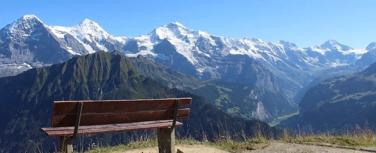 A wooden park bench facing a mountain range with snow-capped peaks and green valleys.