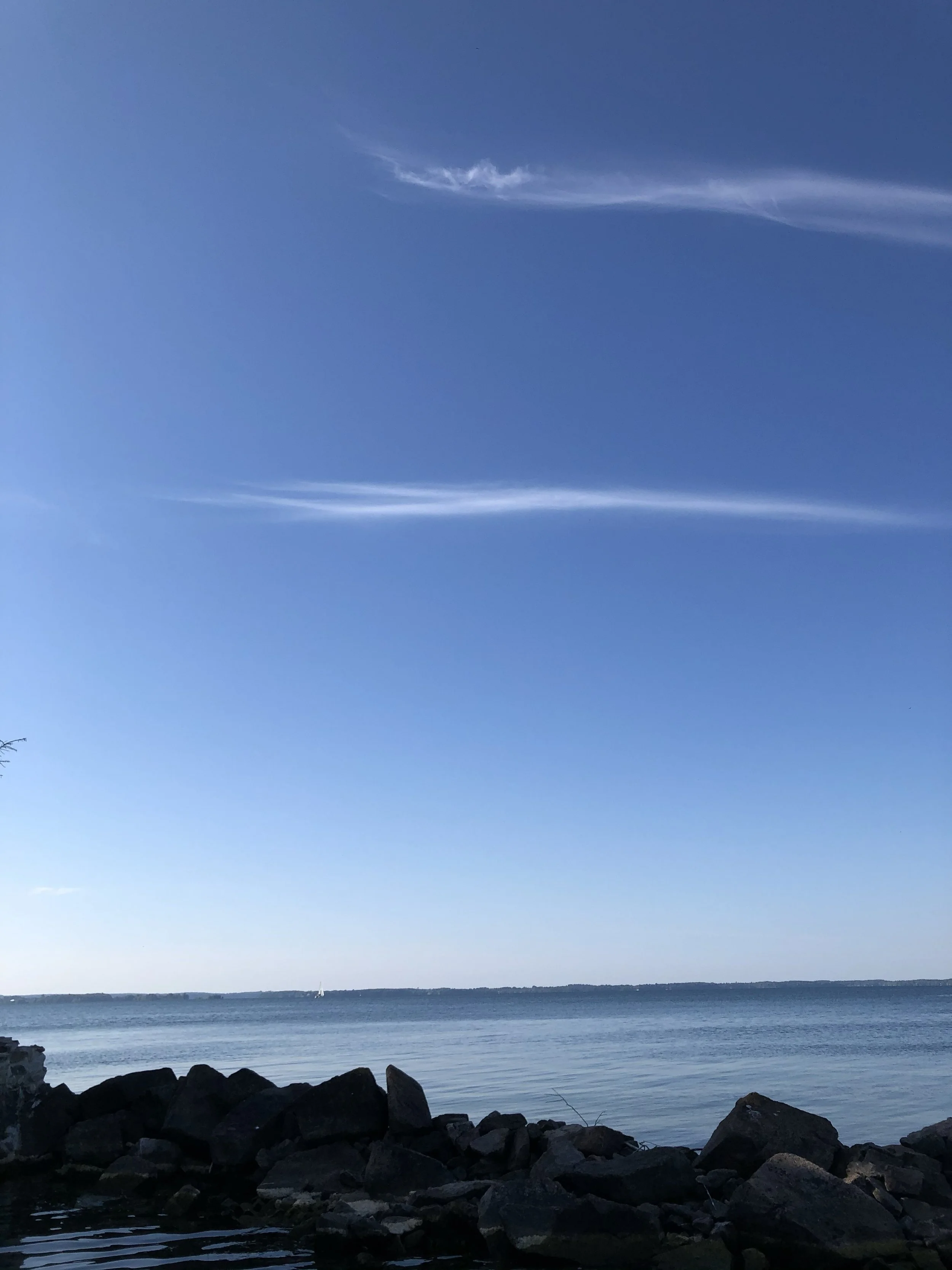 A calm body of water with rocks along the shoreline in the foreground, a distant sailboat, and a clear blue sky with a wispy cloud.
