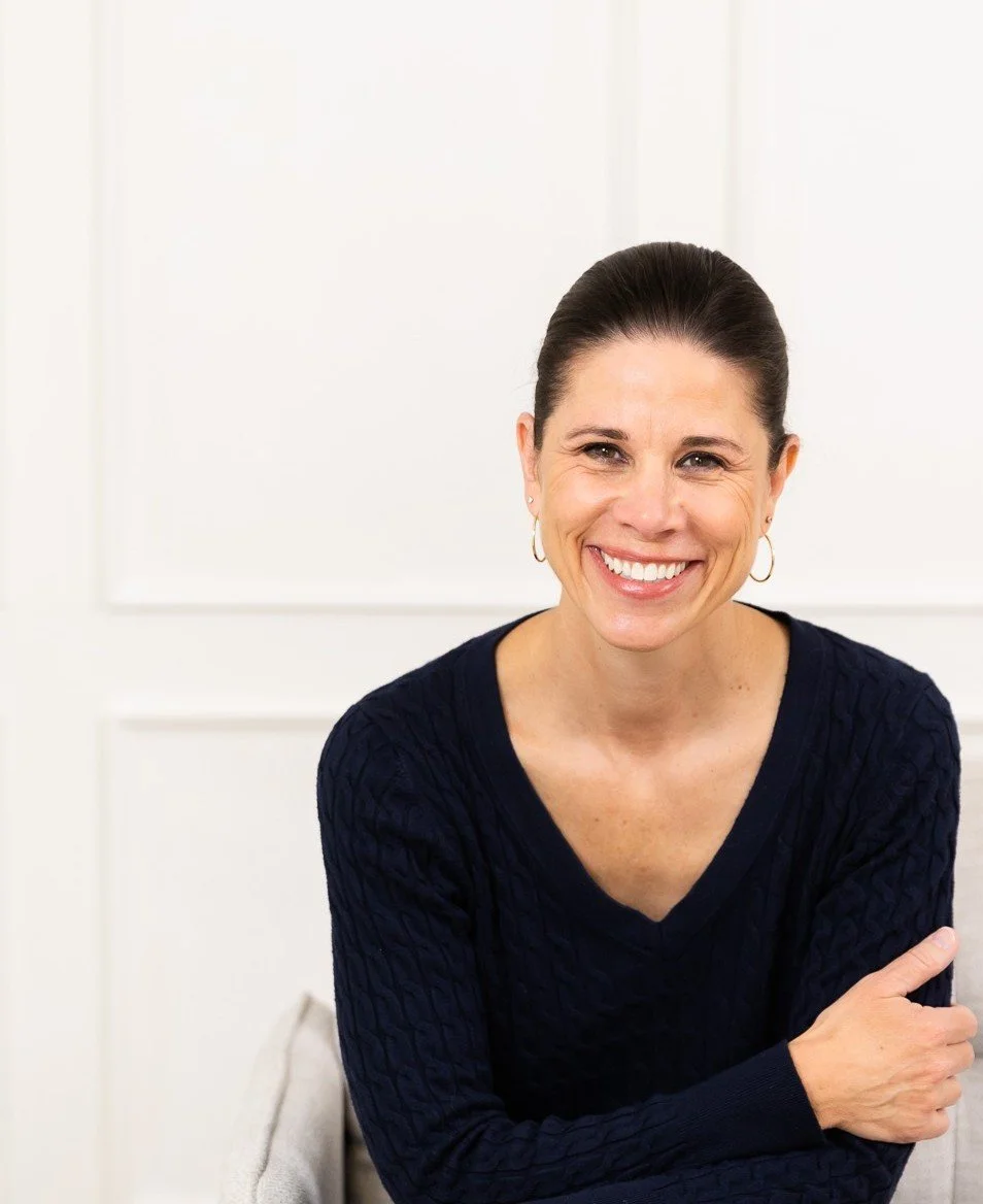 Tanya Melykuty, smiling woman with dark hair pulled back, wearing a navy blue sweater and hoop earrings, sitting and leaning forward, in calm workspace for virtual therapy in Ontario