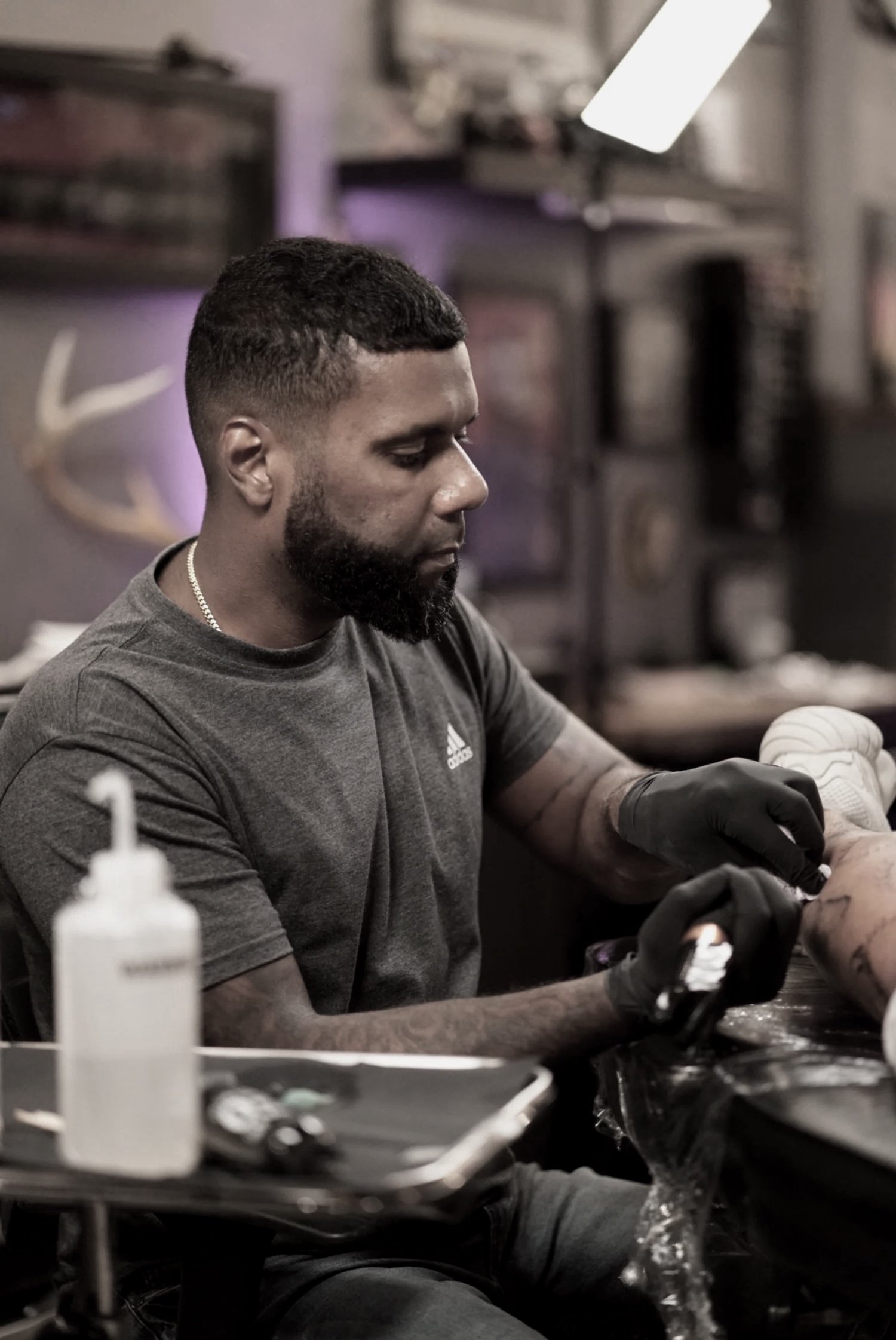 A man with short hair and a beard getting a tattoo on his arm in a tattoo studio. He is wearing a dark gray Adidas t-shirt and black gloves. There are tattoo supplies on a tray on the table in front of him.