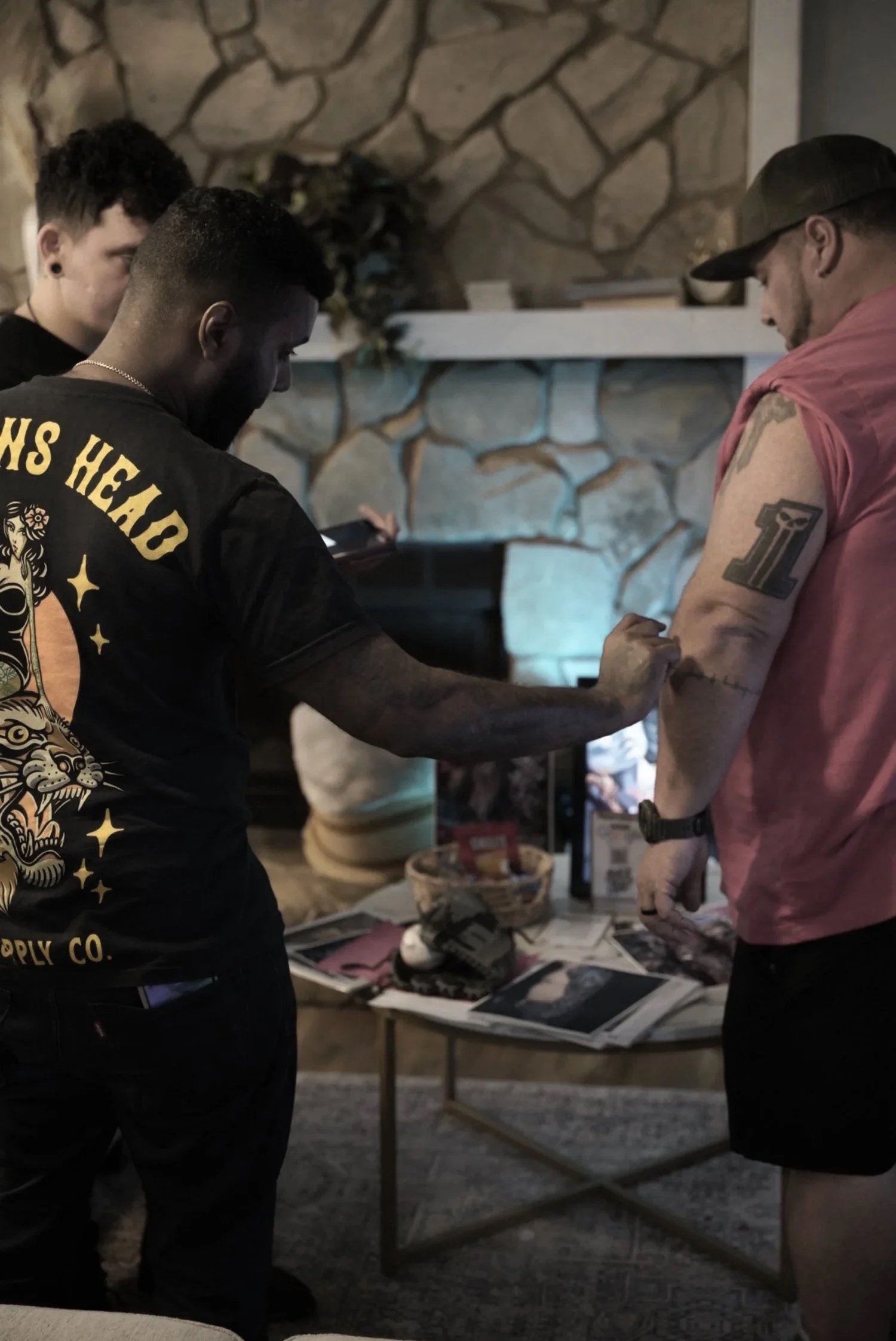 Two men getting tattoos, one with black shirt with tiger and stars print, the other with pink sleeveless shirt, in a room with stone wall, a table with magazines and a TV in the background.