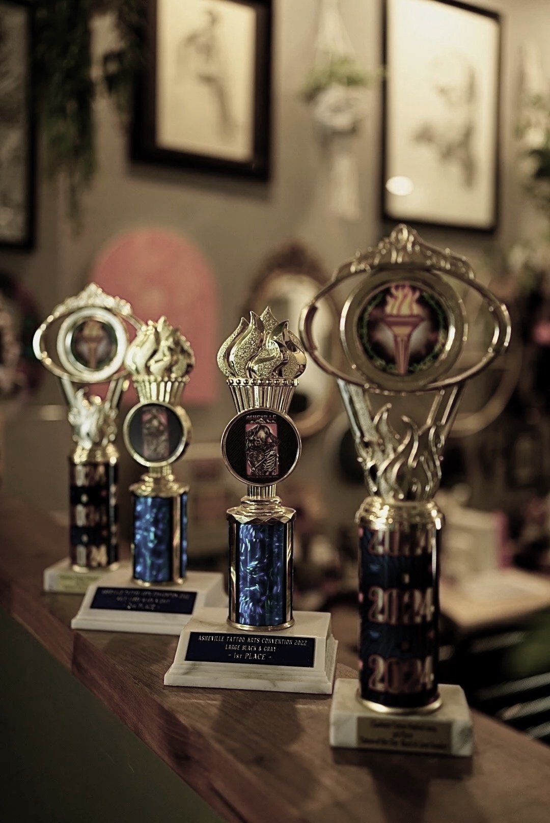 Trophies awarded at the Asheville Tattoo Arts Convention 2022 for large black and gray tattoo categories, placed on a wooden surface with framed artwork on the wall behind.