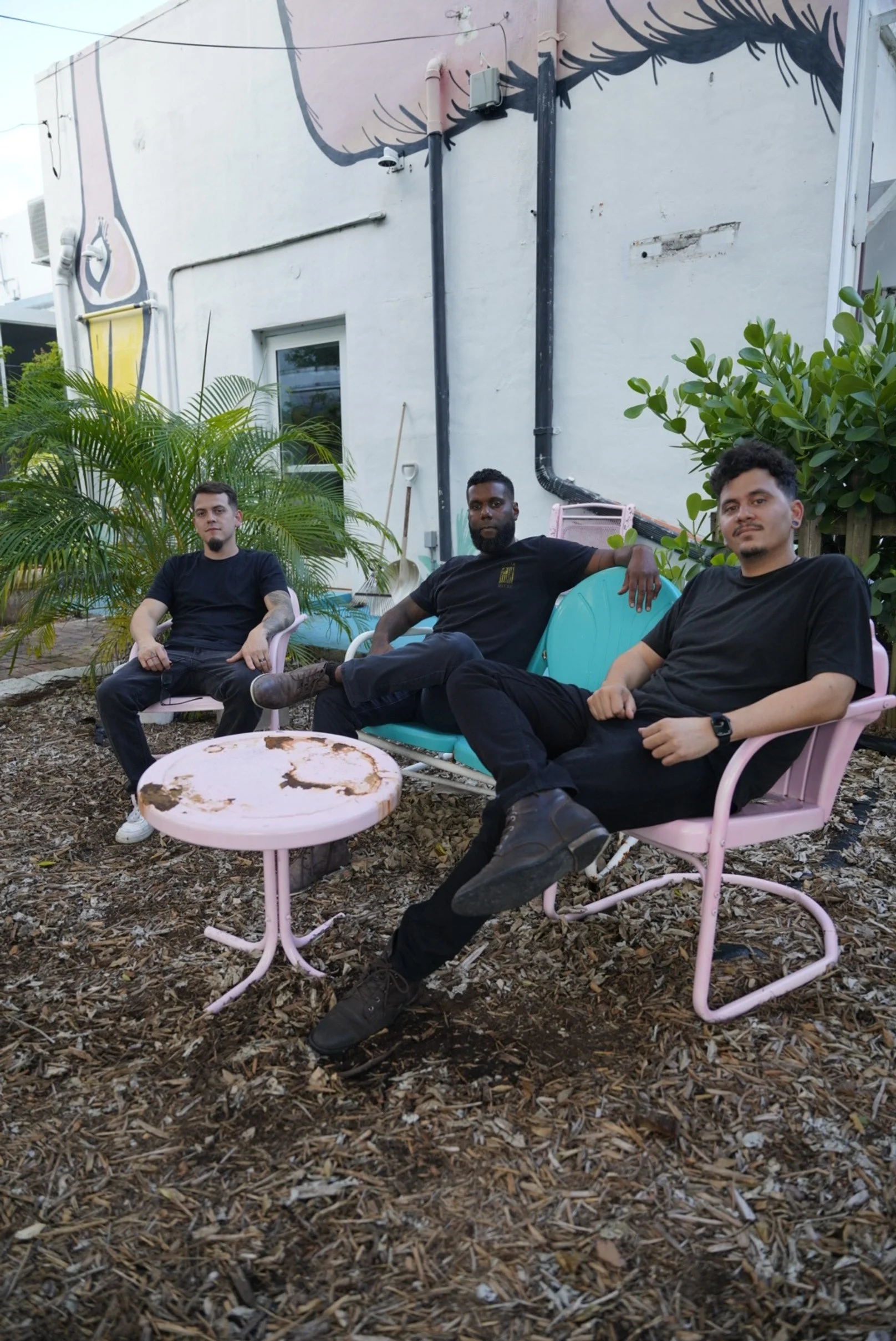 Three men sitting on patio furniture outside near a white wall with black and pink murals, surrounded by plants and mulch, with a rusty pink table in front.