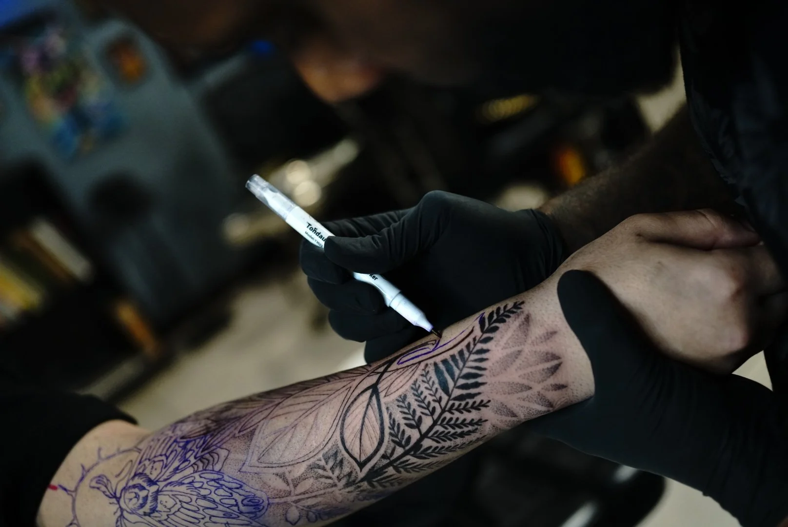 A person getting a tattoo on their forearm, with black gloves on, using a tattoo machine to create the design of leaves and a butterfly.