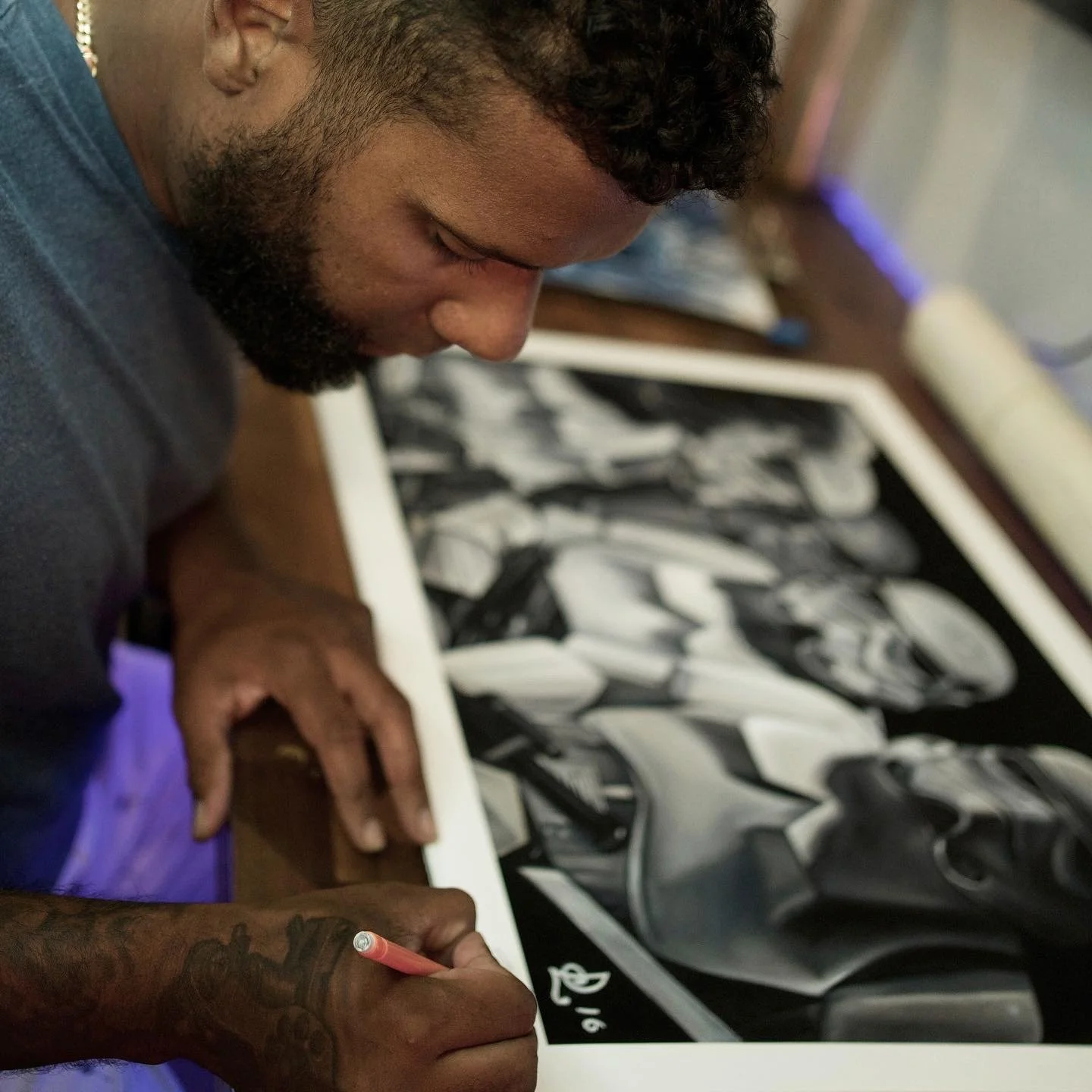 A man with a beard and tattoos on his hand is drawing or signing a black and white painting or illustration on a large table.
