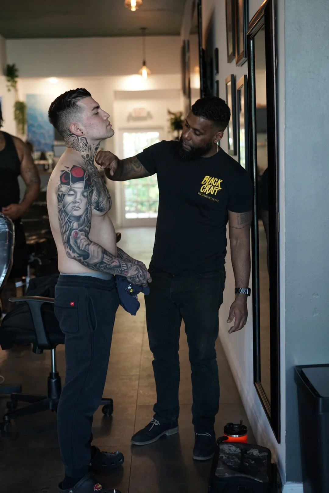 Man with extensive tattoos standing shirtless as tattoo artist prepares to tattoo his chest in a tattoo studio.