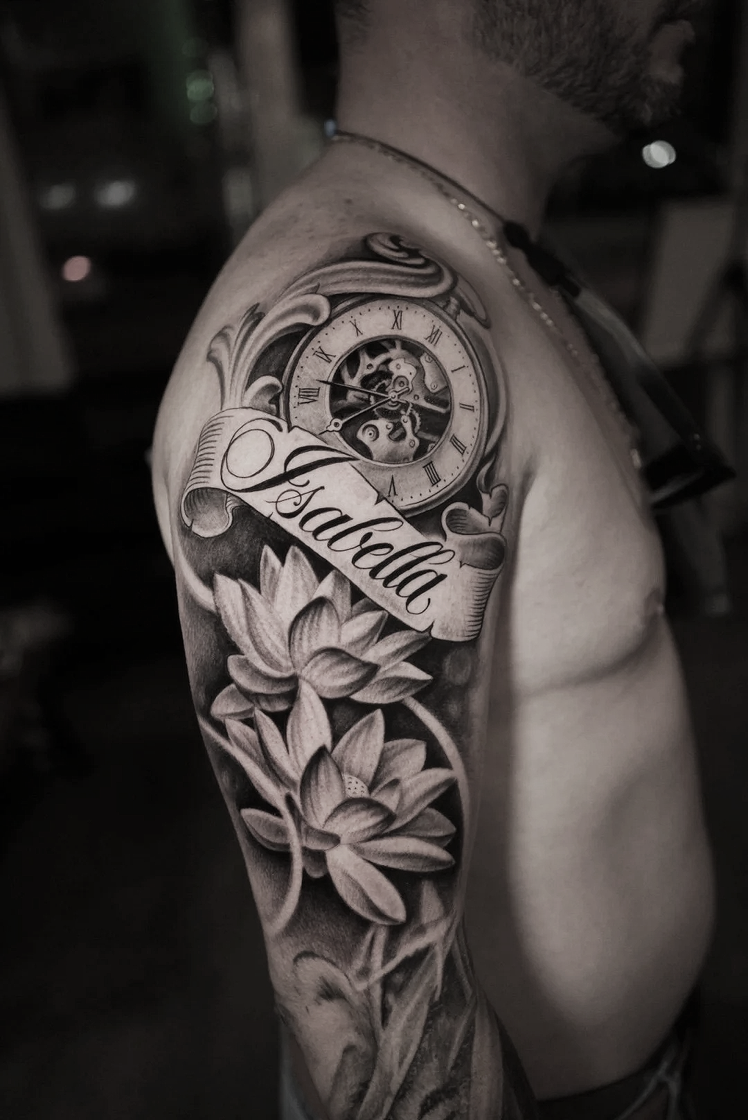 Black and gray tattoo on a person's upper arm featuring a pocket watch, flowers, and the name 'Isabella' on a banner.