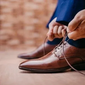 Close-up shot of a man tying the laces of shiny, pointed-toe brown leather dress shoes, wearing dark blue suit pants, against a warm wooden floor background.