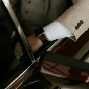 Close-up of a man's forearm and wrist, wearing a modern smartwatch with a brown leather strap, visible beneath a beige linen jacket sleeve with three brown buttons.