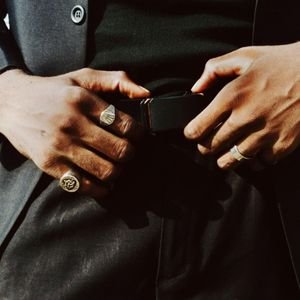 Close-up of a man's hands with rings buckling a black leather belt over dark trousers, emphasizing the detail of his attire.