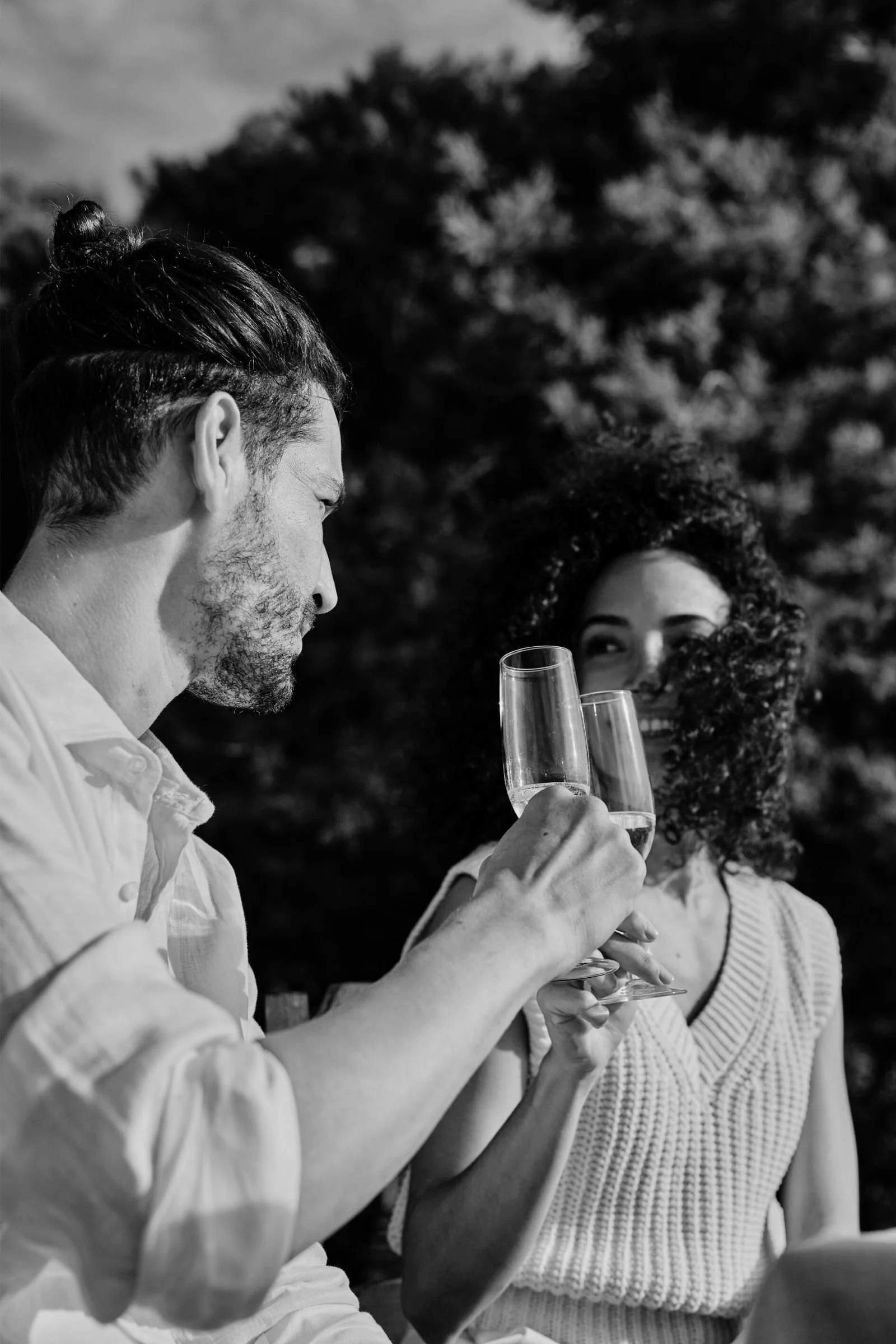 Black and white close-up of a stylish couple toasting with champagne flutes outdoors, focused on the man in a linen shirt.