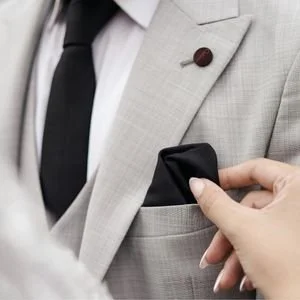 Close-up of a woman's hand adjusting a folded black silk pocket square in the chest pocket of a man's light grey suit jacket.