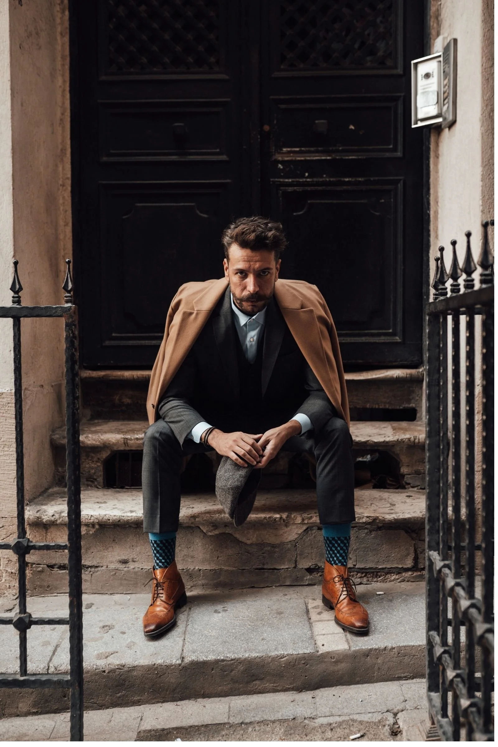 Stylish man sitting on urban steps, wearing a dark suit, tan coat draped over his shoulders, and patterned blue socks with brown boots.