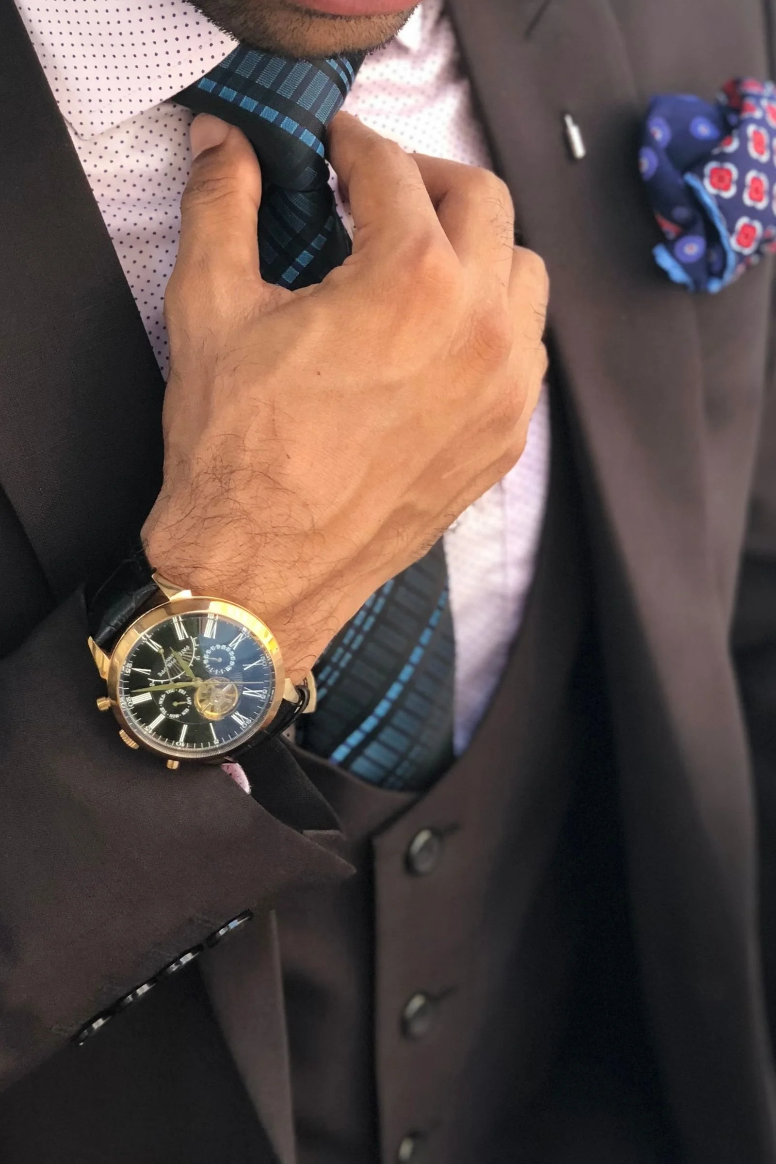 Close-up of a man adjusting a striped tie, wearing a dark brown suit vest and a gold watch, with a colorful pocket square visible in his jacket pocket.
