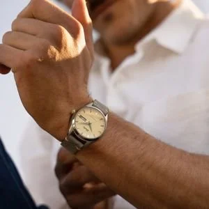 Close-up of a man's wrist outdoors in sunlight, wearing a classic analog watch with a light face and stainless steel mesh band, paired with a casual white button-down shirt.