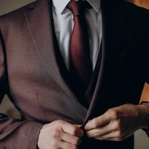 Detailed shot of a man fastening the button of a textured, deep burgundy suit jacket, worn over a matching waistcoat, white shirt, and a rich maroon tie.