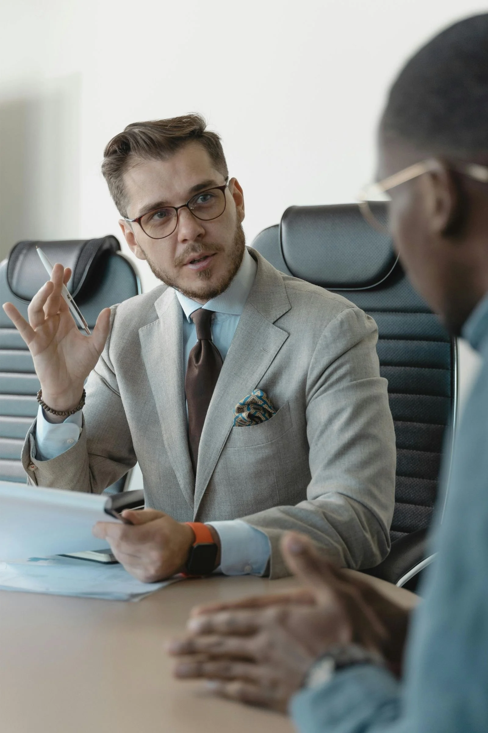 Professional man in a light tan suit and glasses gesturing while discussing documents with a client across a table.