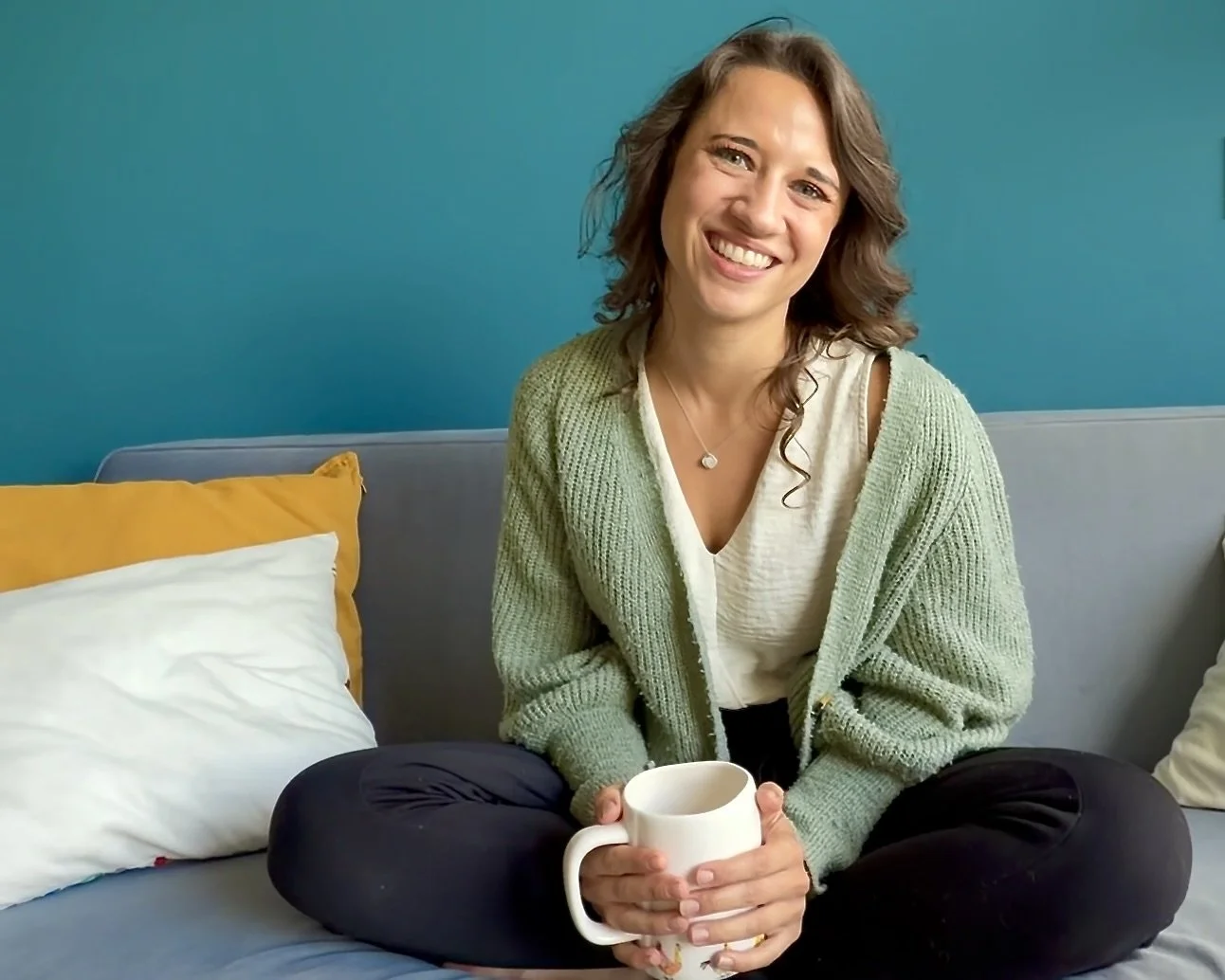 A woman with curly brown hair sitting on a gray couch with colorful pillows, holding a white mug, smiling at the camera, against a blue wall.