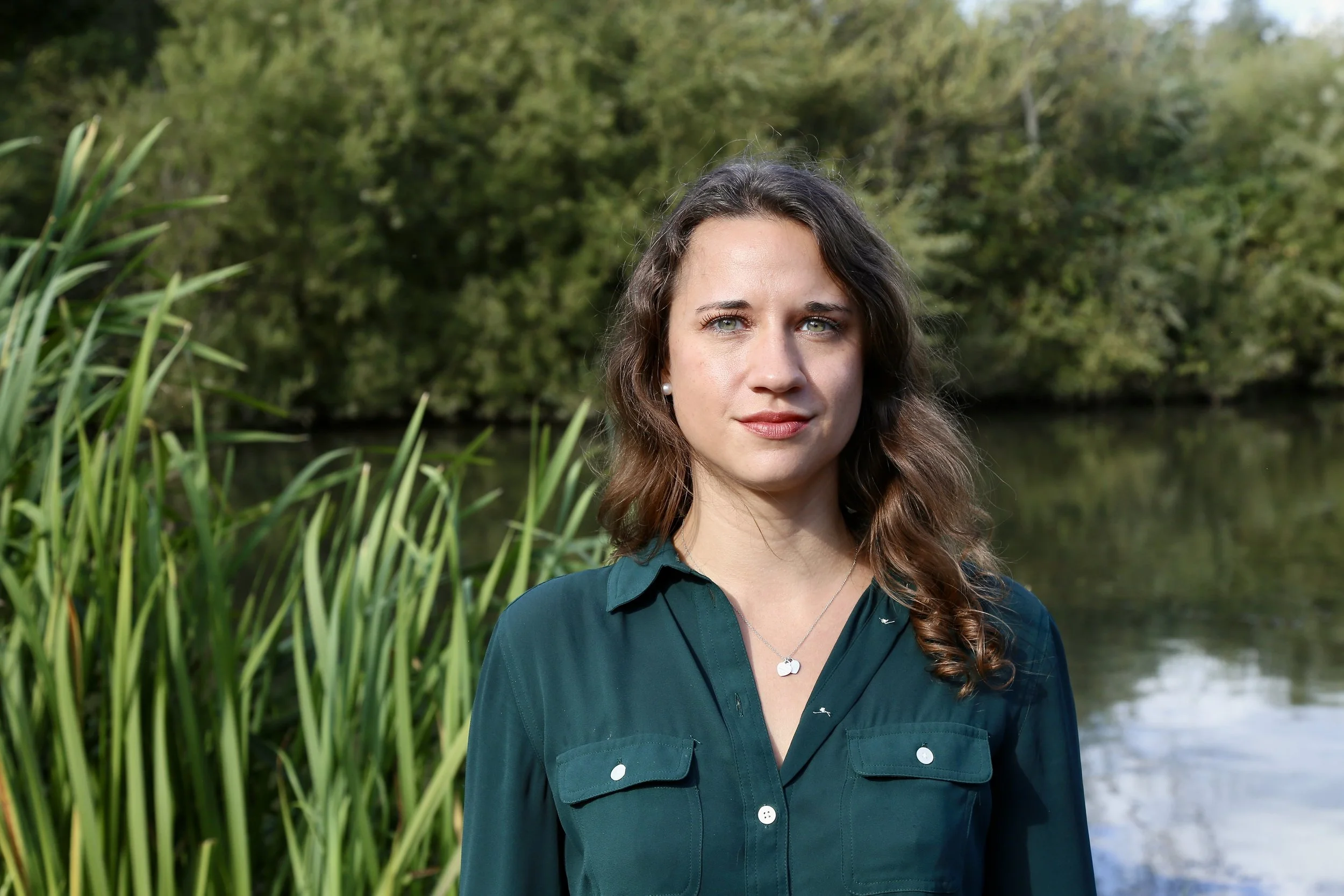 A woman standing outdoors near a body of water, surrounded by trees and tall grass.