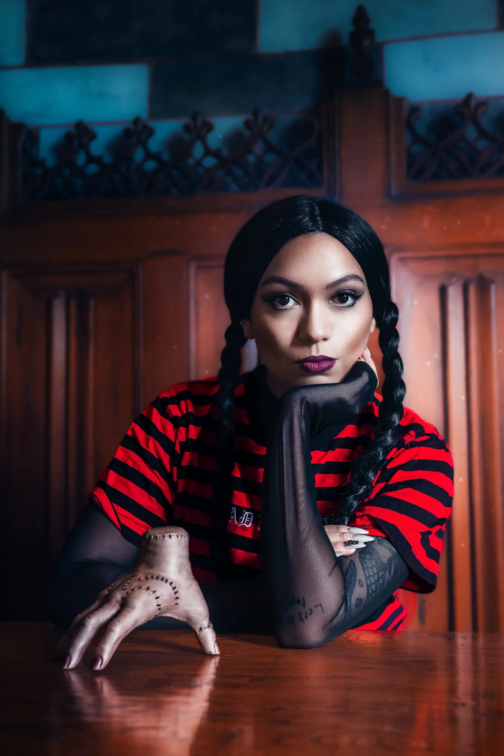 Une femme avec des cheveux noirs tressés, portant un t-shirt rouge à rayures noires et des gants noirs, posant dans une pièce avec un fond en bois, regardant fixement l'appareil photo.