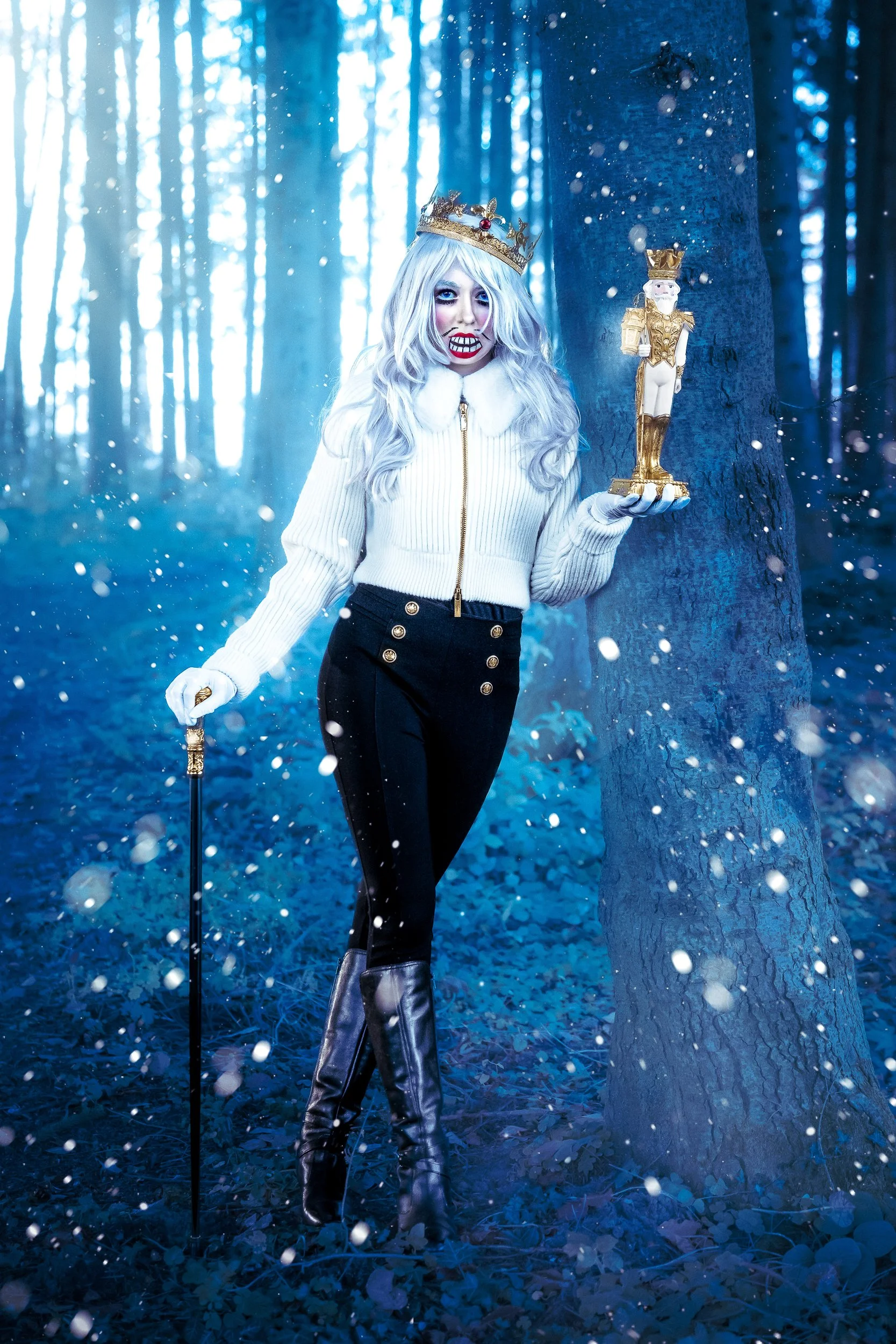 Une femme déguisée en reine avec une couronne dorée, une robe blanche et des bottes noires, tenant une petite statue dorée et un bâton, dans une forêt enneigée avec des flocons de neige tombant.