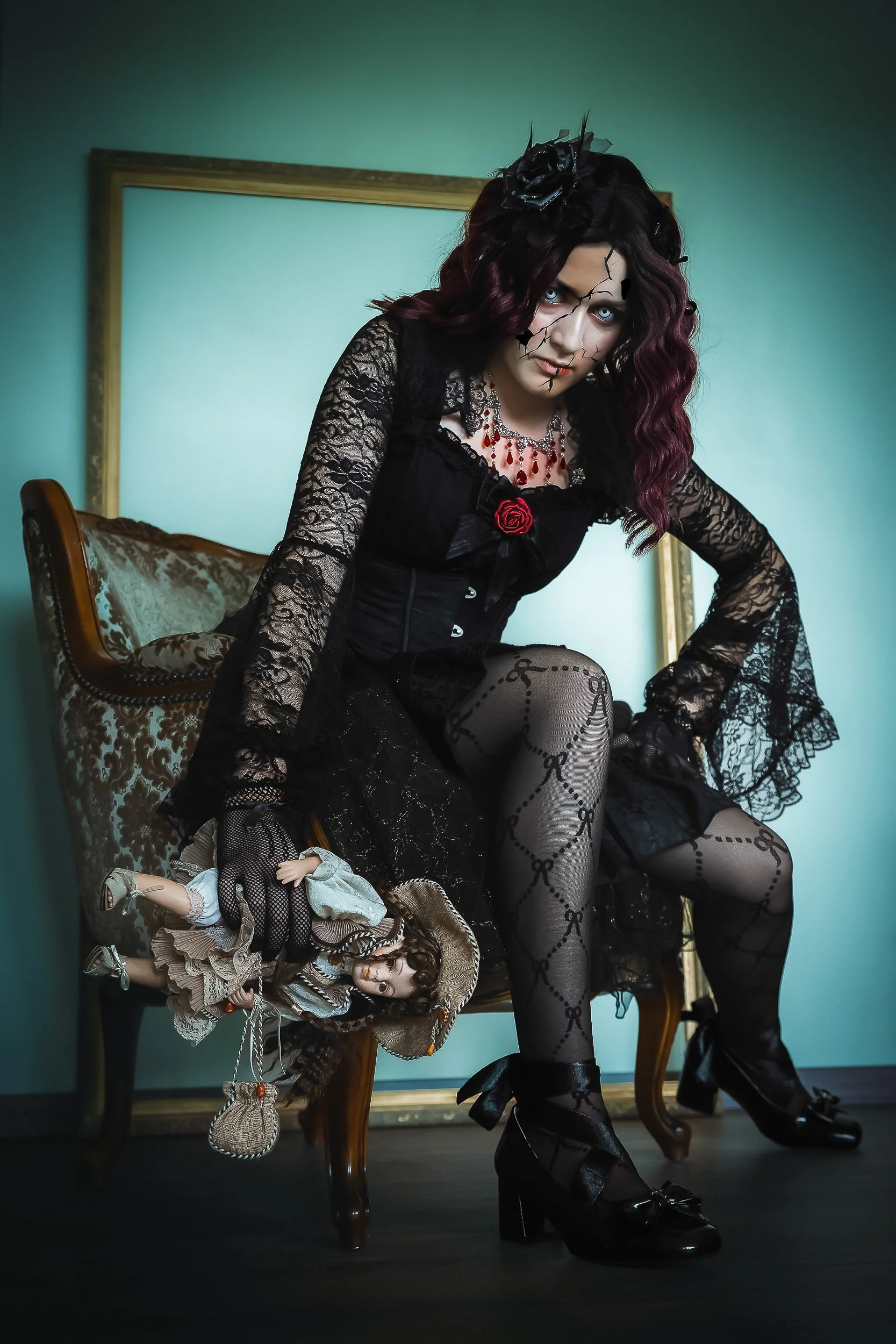 Une femme vêtue en style gothique, portant une robe noire en dentelle, des collants avec un motif en chaîne, des chaussures à talons et un collier rouge. Elle est assise sur un fauteuil ancien, tenant une poupée habillée en costume historique, avec u