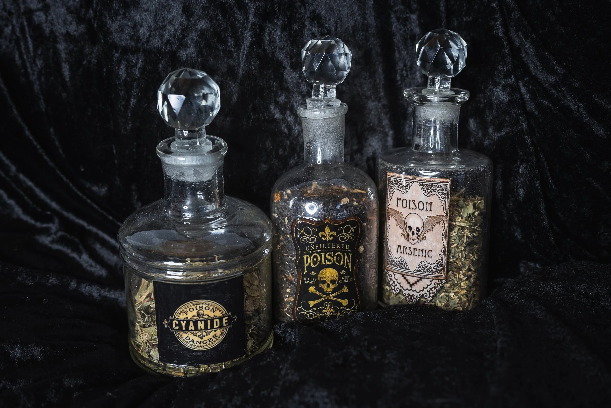 Trois fioles en verre contenant des herbes, étiquetées comme poison, poison non filtré et arsenic, sur un fond de tissu noir.