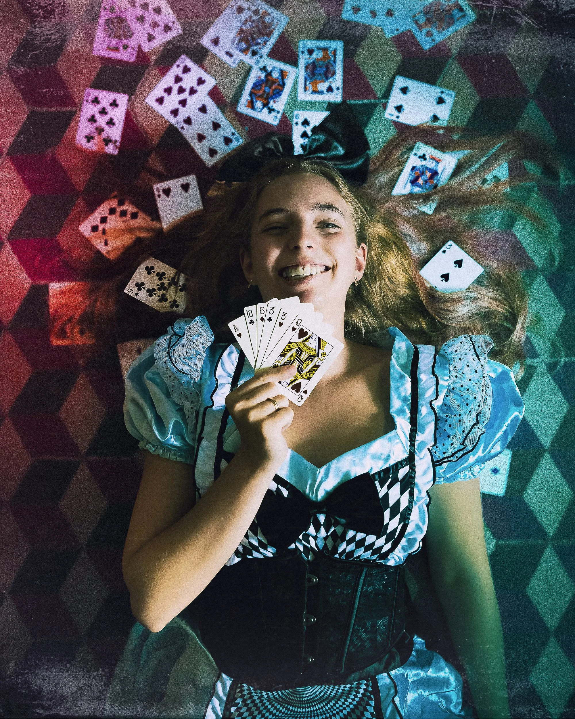 Une jeune femme habillée en Alice au Pays des Merveilles, allongée sur un sol en damier, entourée de cartes à jouer, tenant un éventail de cartes à jouer dans la main.