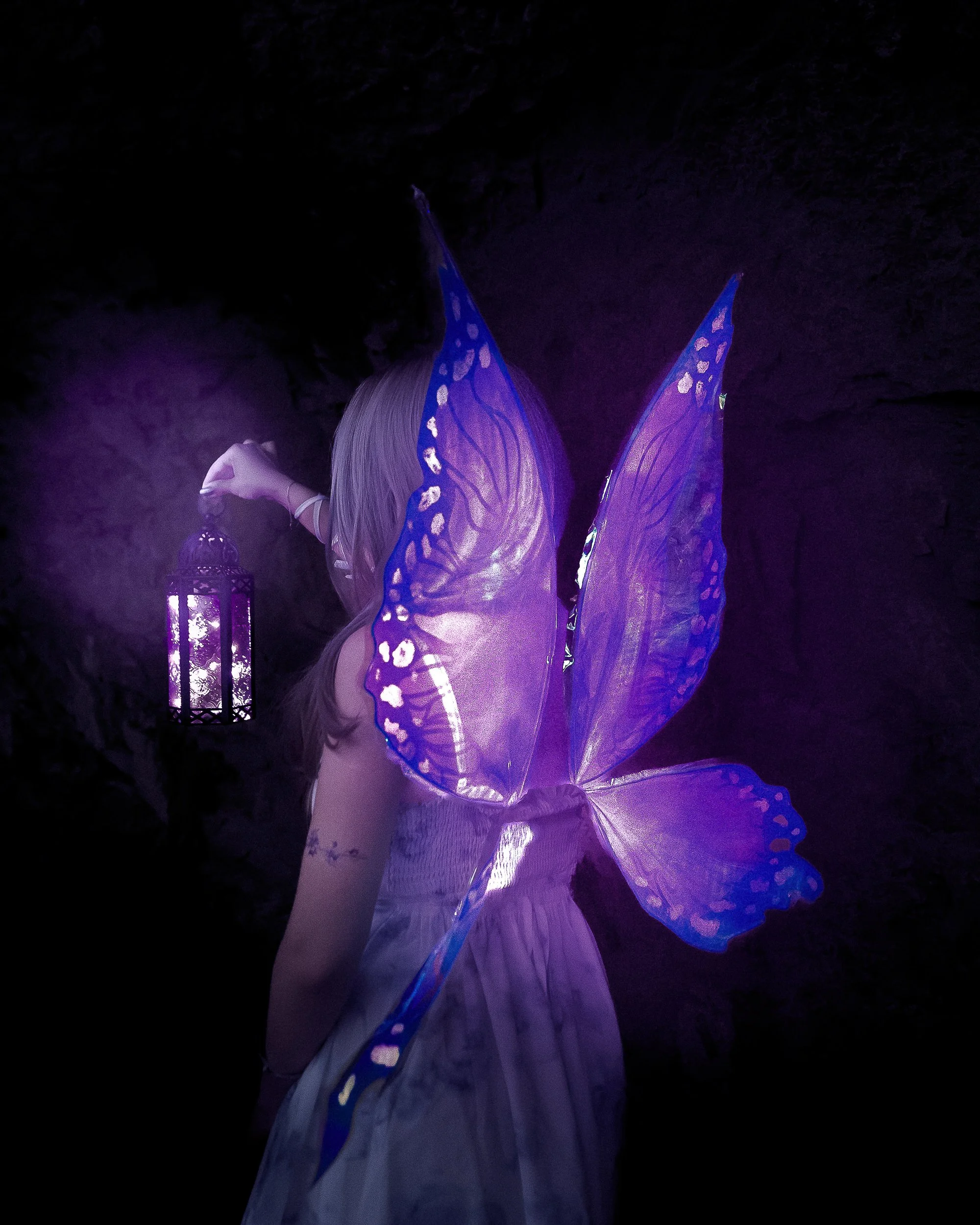 Fée avec des ailes violettes translucides, tenant une lanterne violette dans un endroit sombre.