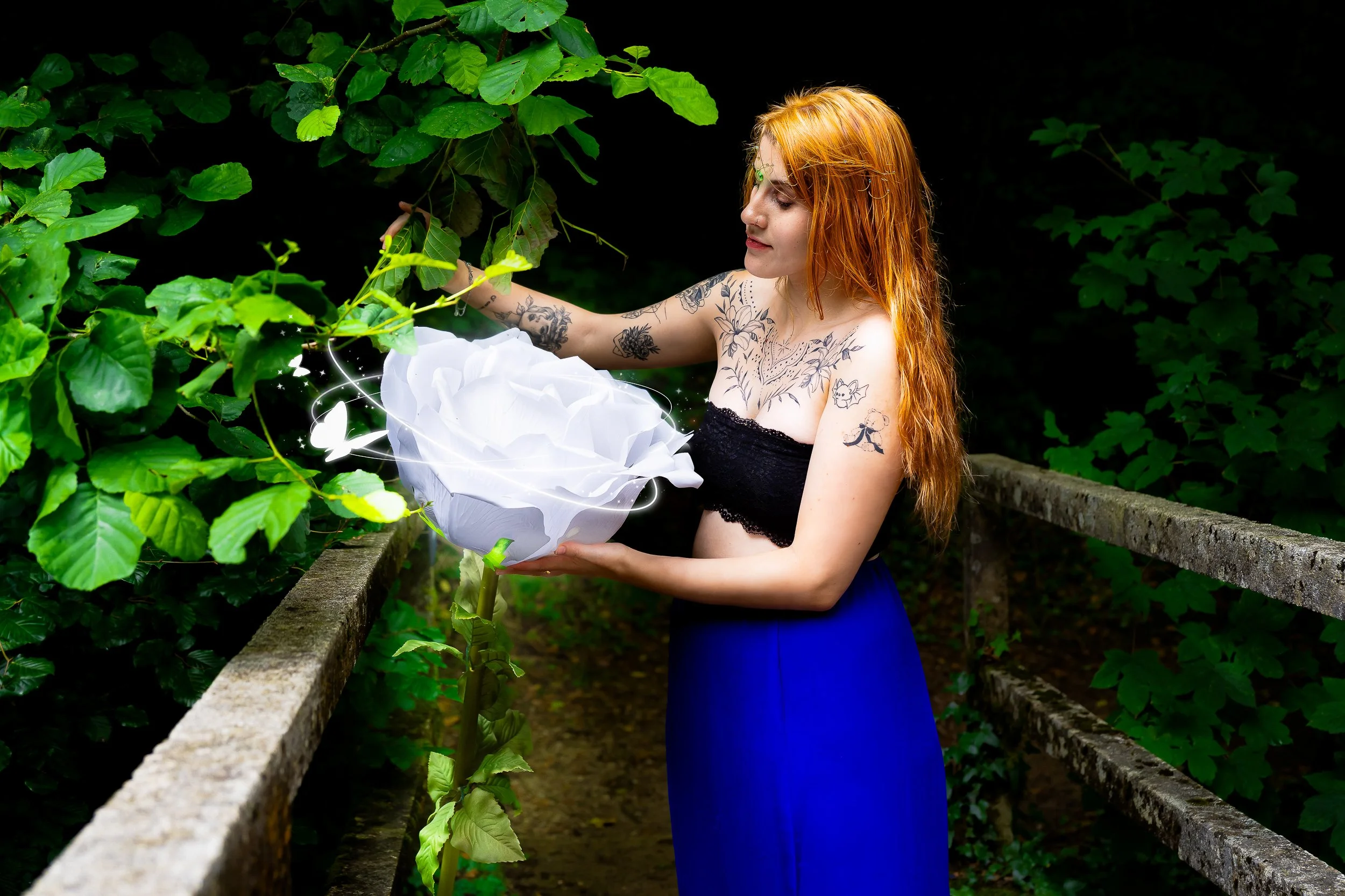 Jeune femme aux longs cheveux roux, tatouages, portant un haut noir et une longue jupe bleue, tenant une grande fleur blanche en papier dans un environnement naturel vert, sur un pont en pierre.