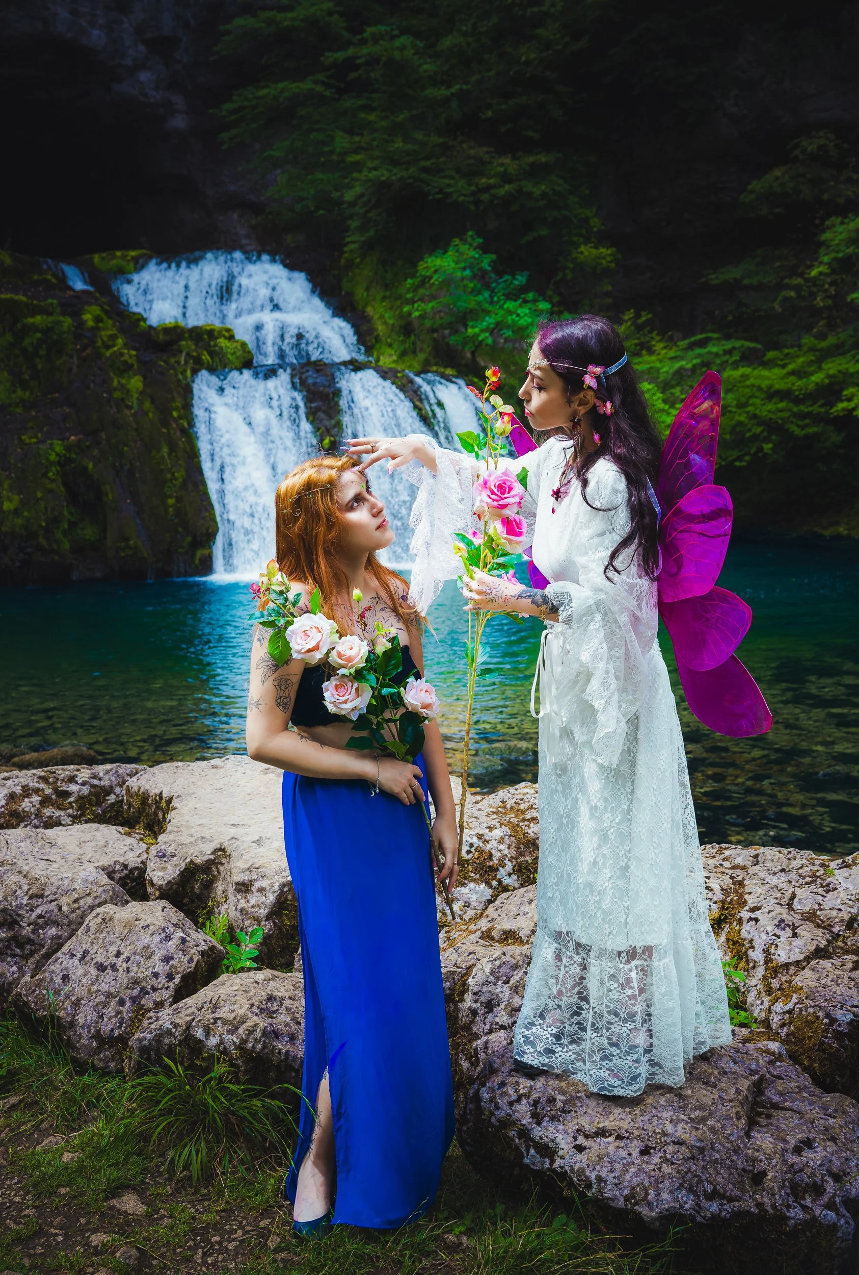 Une scène féerique avec deux femmes, l'une habillée en fée avec des ailes violettes, tenant un bouquet de roses, et l'autre portant une robe bleue, dans un décor naturel avec une cascade et une rivière en arrière-plan.