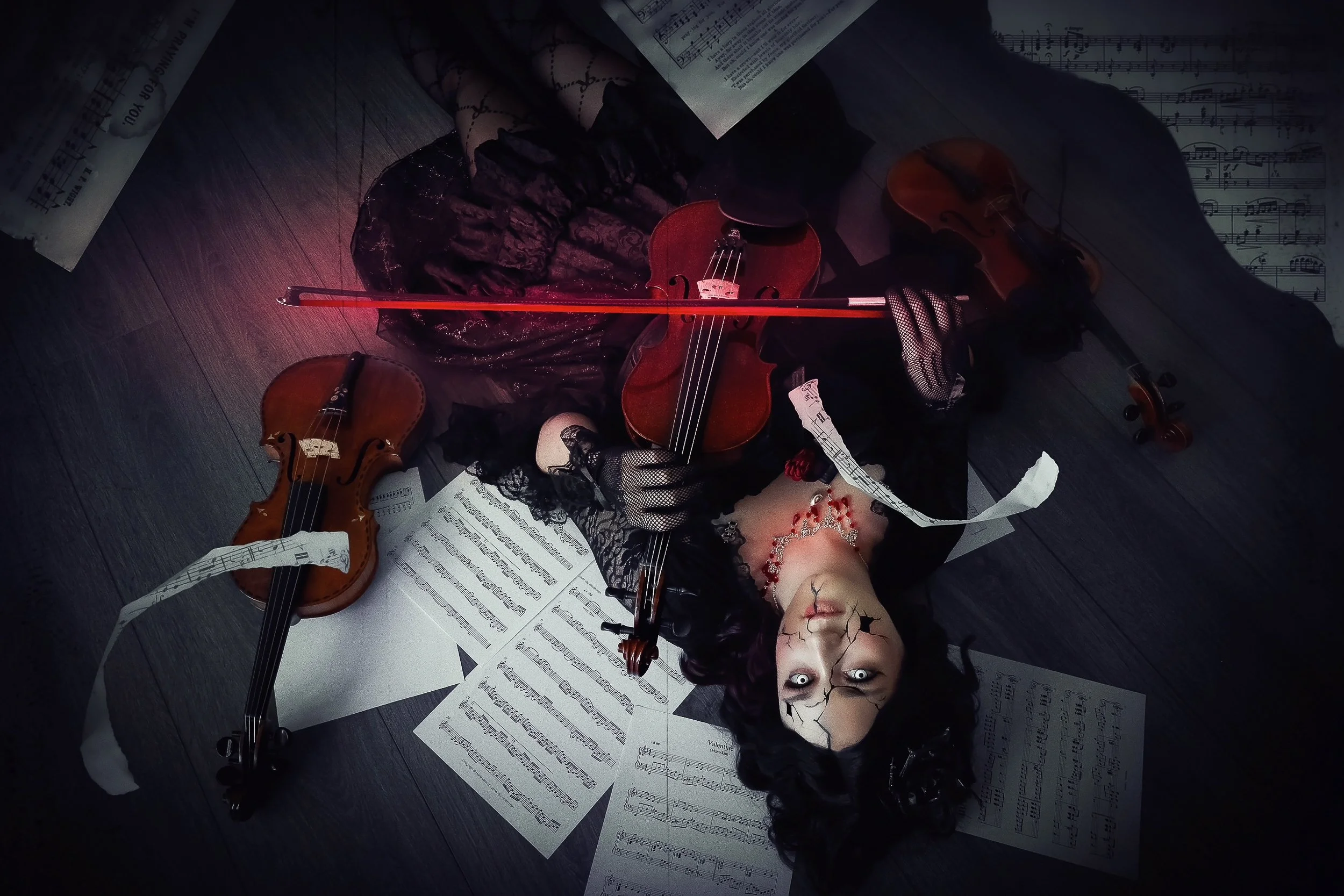 Une femme déguisée en personnage gothique avec des yeux pâles, des tatouages faciaux, portant une robe noire, qui tient un violon et un archet rouge, entourée de partitions de musique dispersées sur le sol en bois.