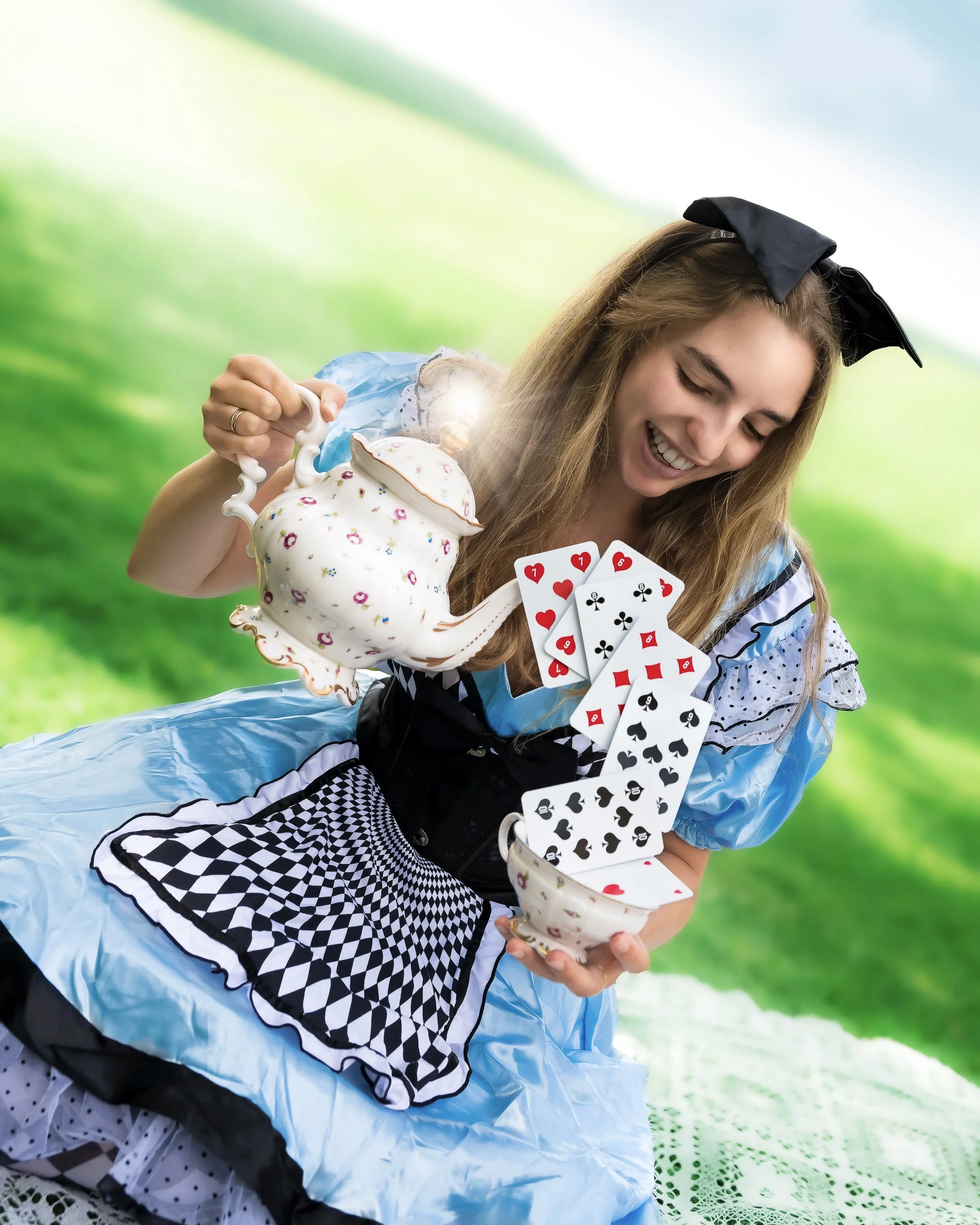 Une femme habillée en Alice au pays des merveilles, souriante, tenant une théière en porcelaine décorative et des cartes à jouer suspendues dans l'air, dans un décor de nature verte.