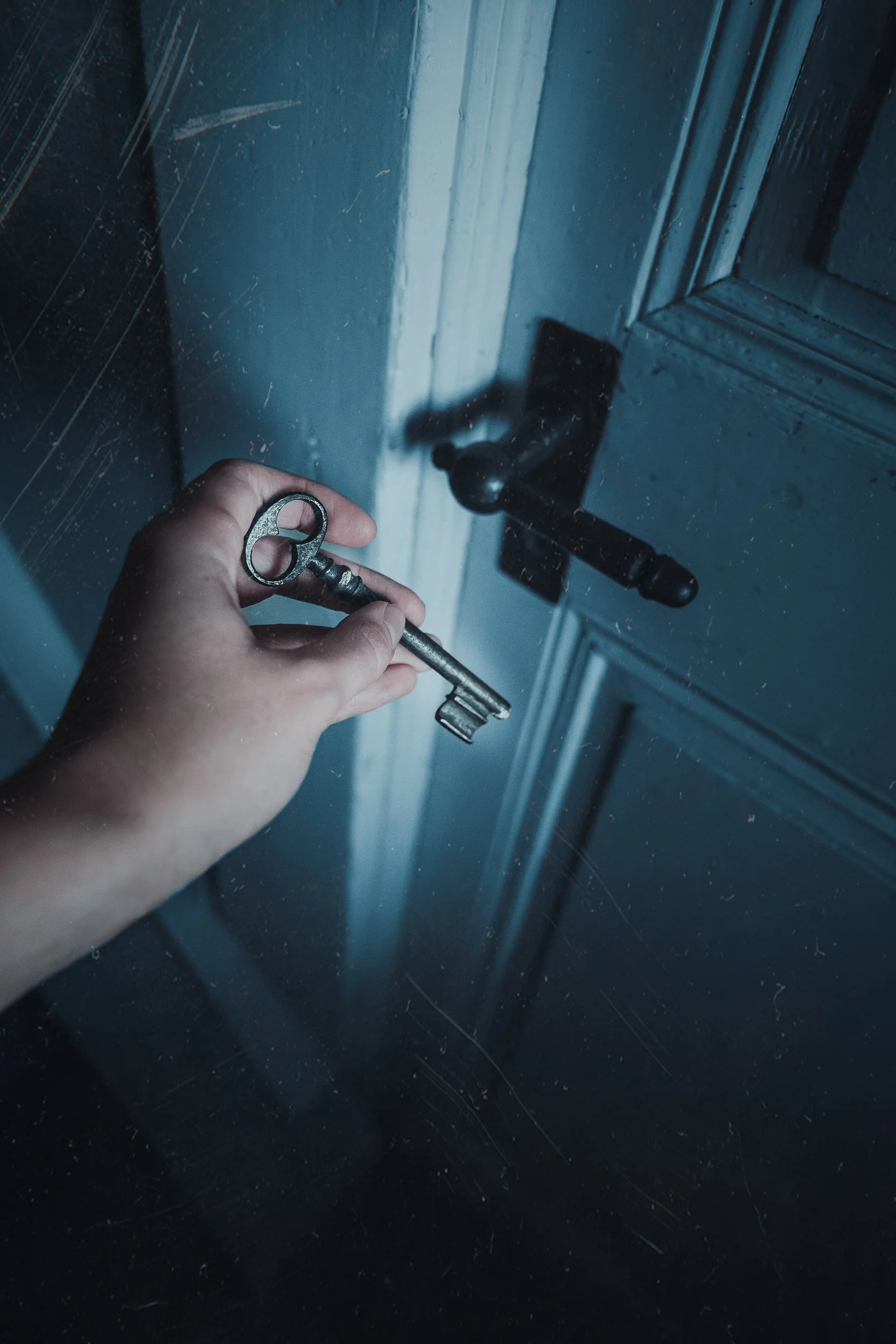 Une main tenant une vieille clé en fer, prêt à ouvrir une porte en bois avec une poignée en métal, dans une pièce sombre.