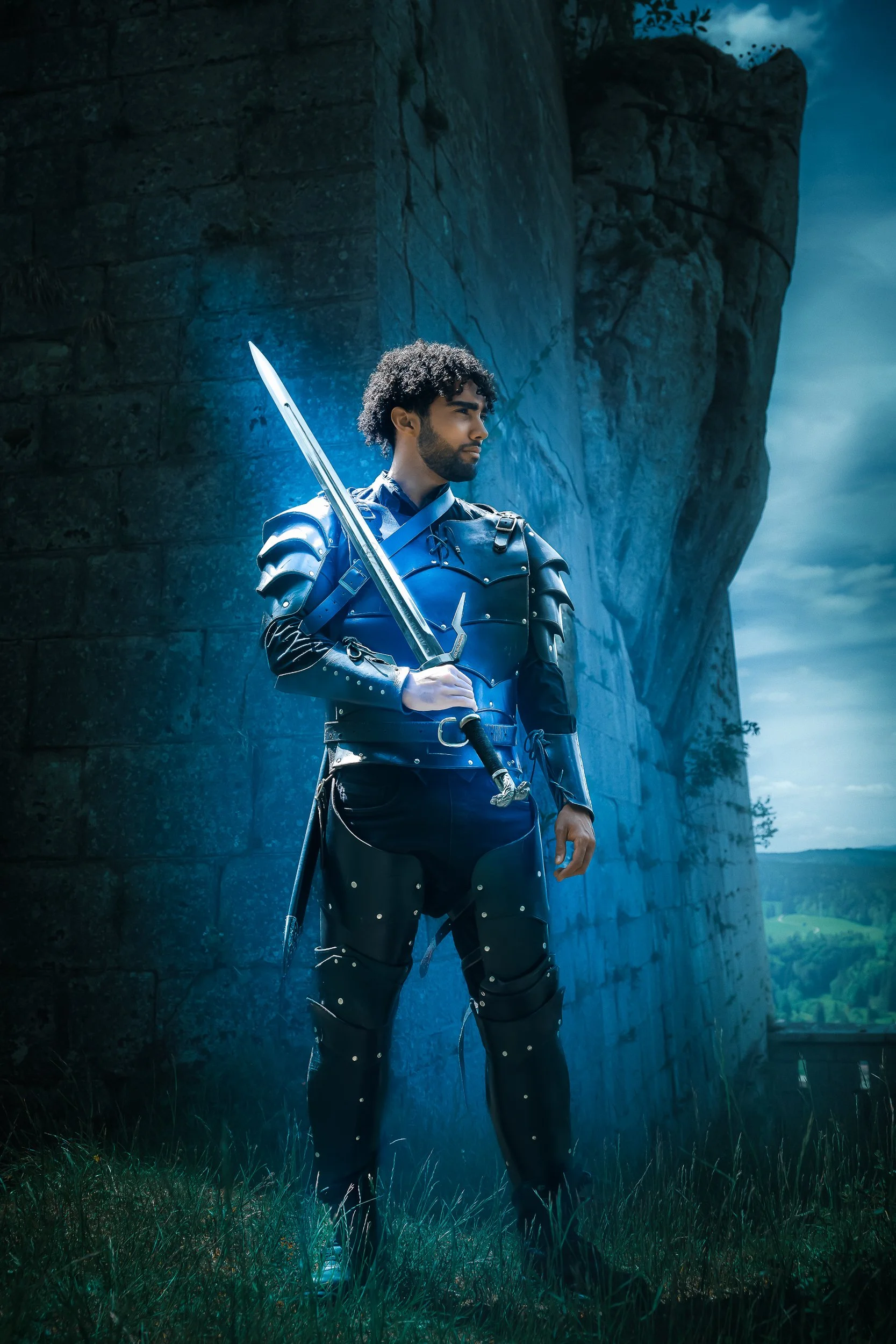 Homme en armure noire, tenant une épée, debout devant un mur en pierre, dans un paysage pastoral.