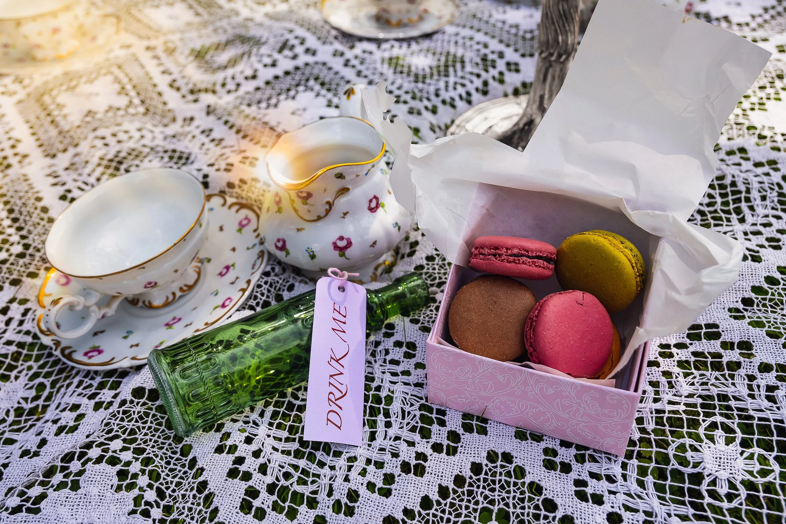 Une caisse à macarons roses, jaunes, et bruns sur une nappe en dentelle, avec une théière et des tasses en porcelaine ornées de fleurs, une bouteille verte portant une étiquette 'DRINK COKE'.