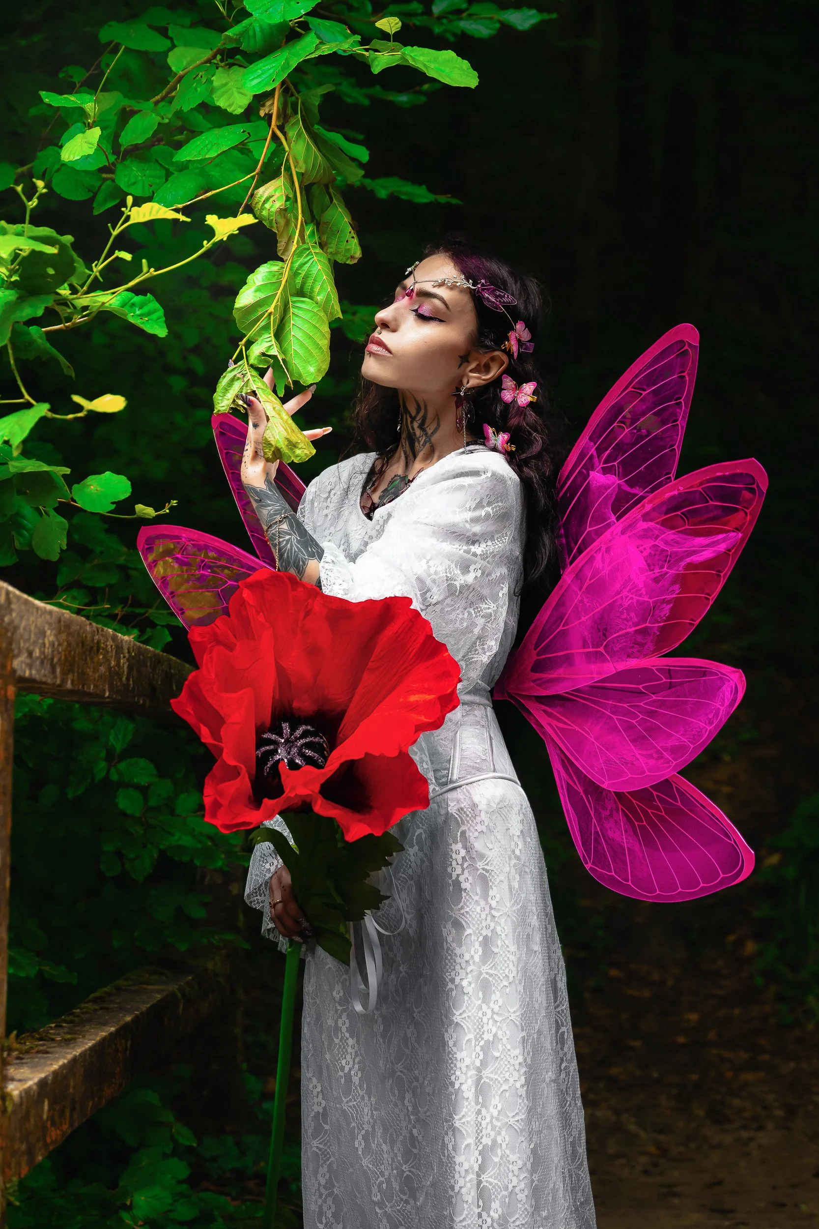 Une femme avec des ailes de fée roses, tenant une grande fleur rouge, en tenue blanche, dans un environnement naturel vert avec des feuilles.
