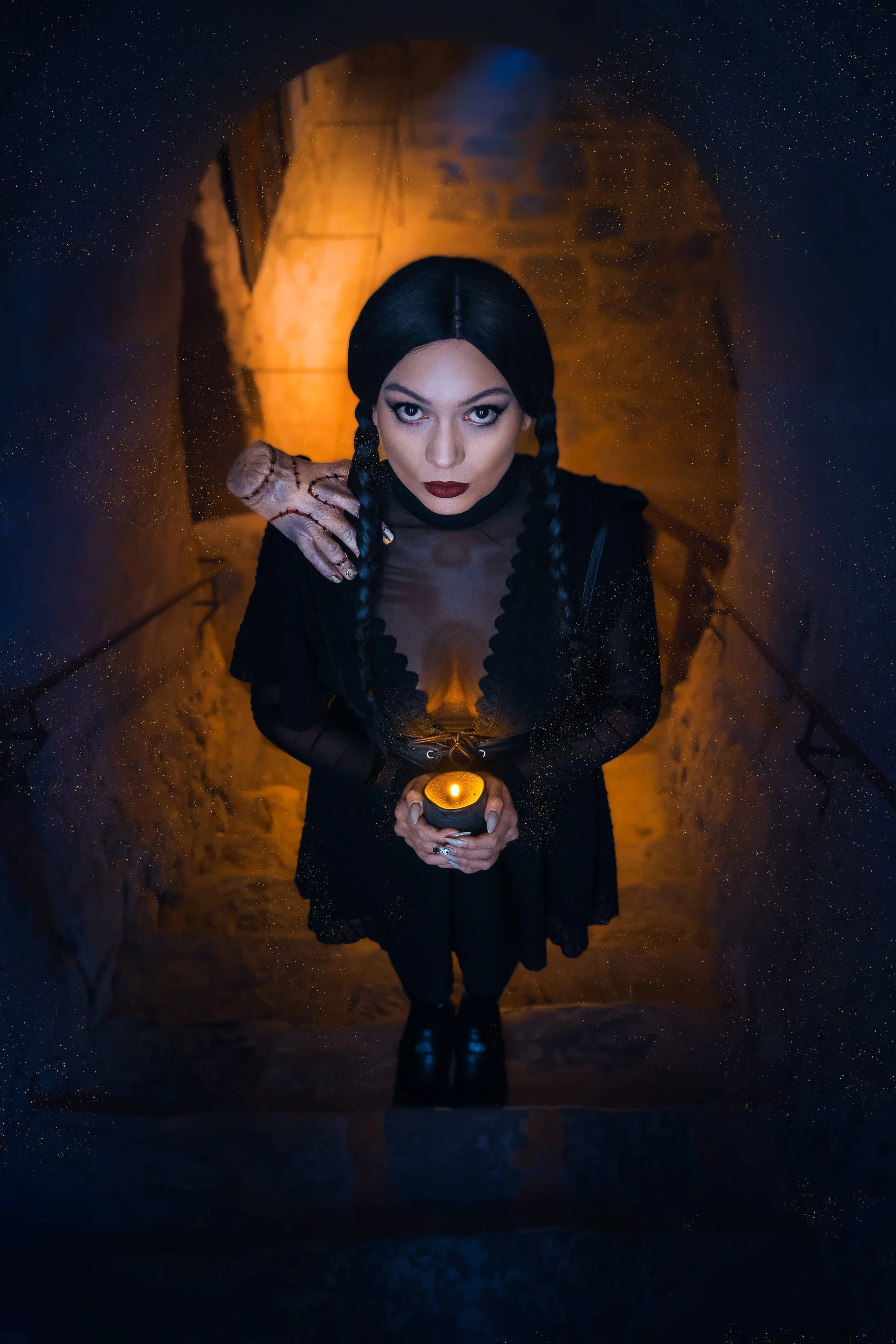 Une femme avec des cheveux noirs tressés, portant un vêtement noir, tenant une bougie orange en pierre, se tenant dans une grotte éclairée d'une lumière chaude.