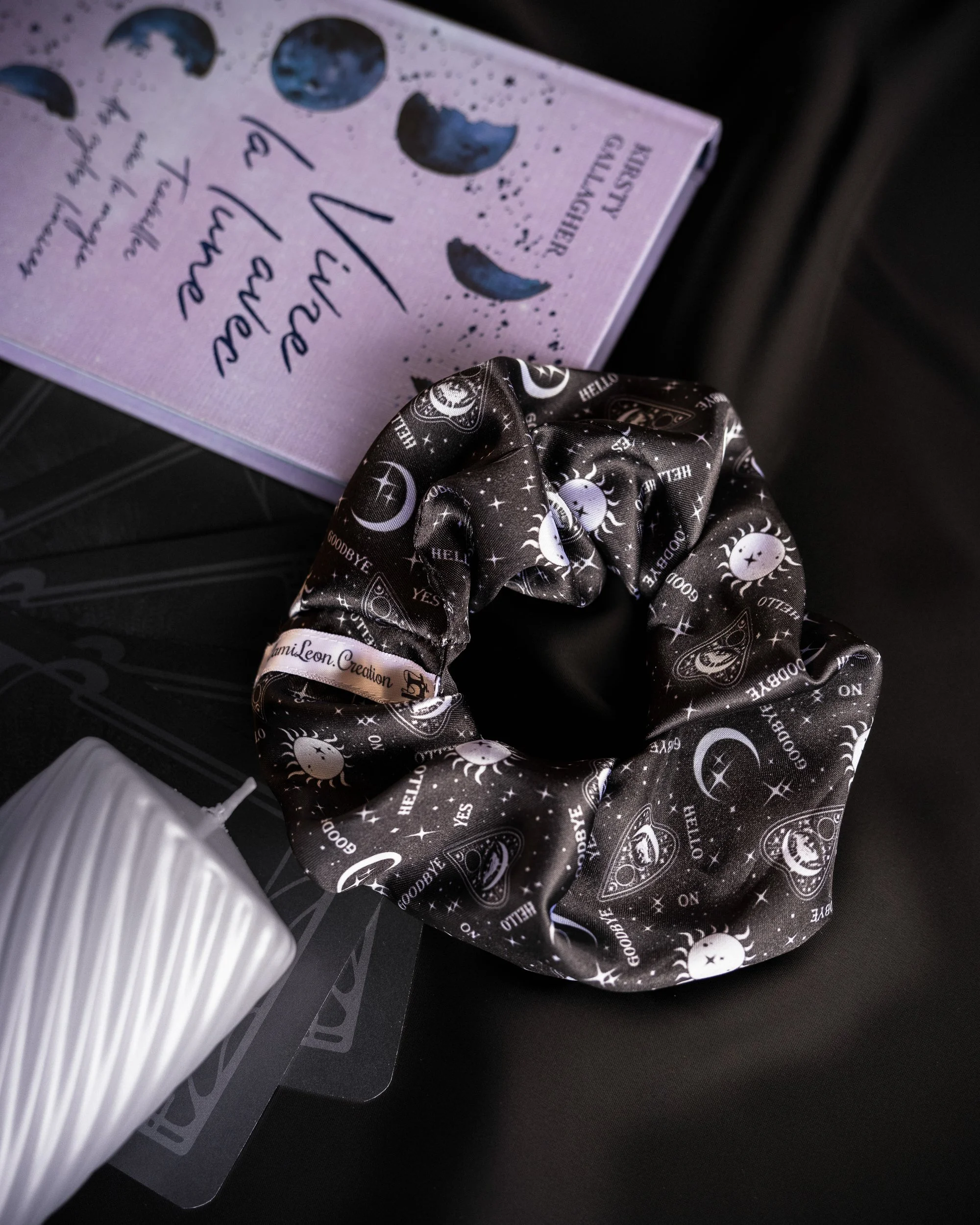 Un foulard noir à motifs d'astres et de lunes, une boîte à livre lilas avec des représentations de lunes et de texte, une bougie blanche à rayures.