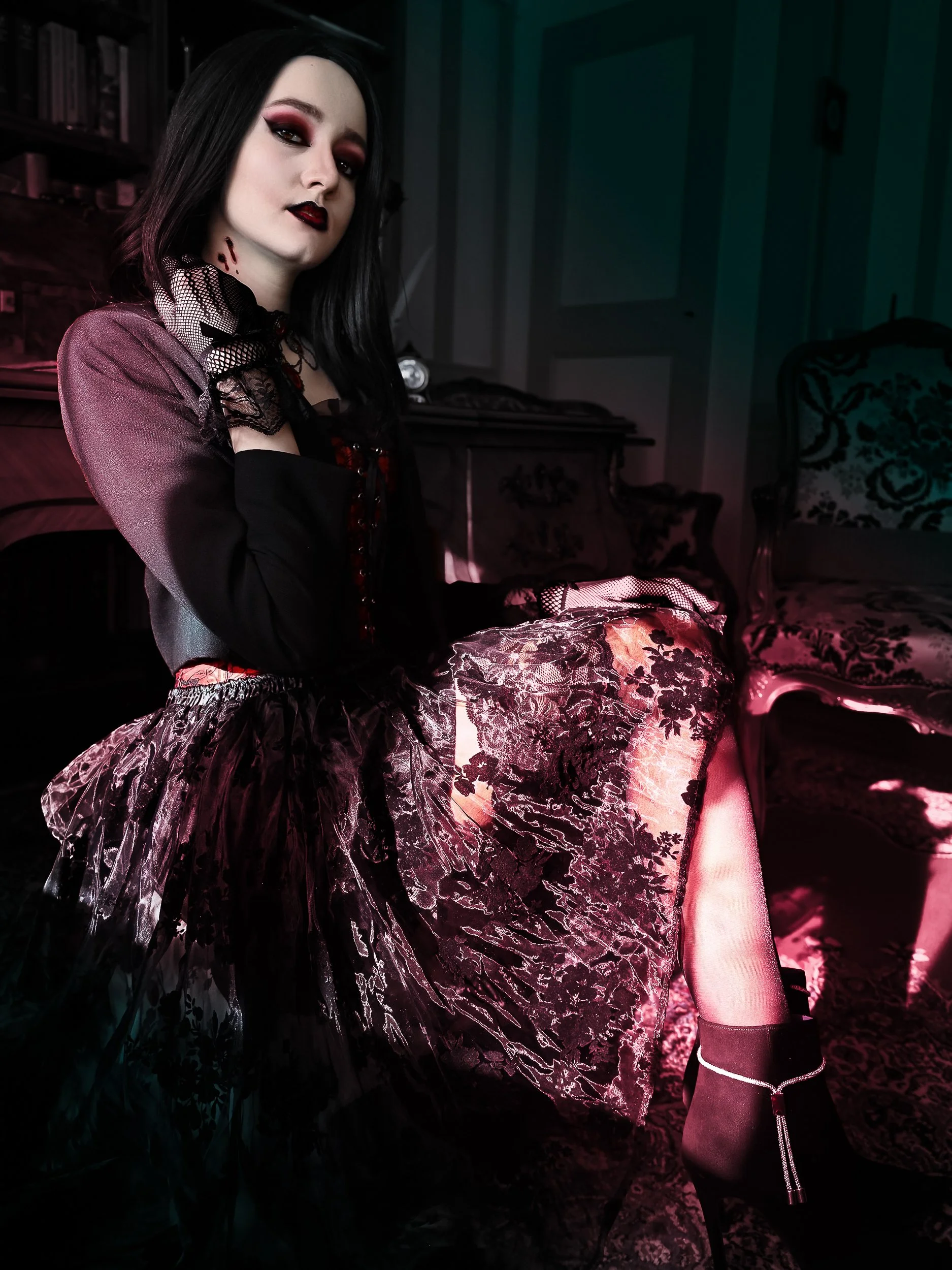 Une femme avec un maquillage sombre, portant une robe en dentelle noire et un accessoire en filet, assise dans une pièce décorée de meubles anciens et de tapis, éclairée par une lumière colorée.