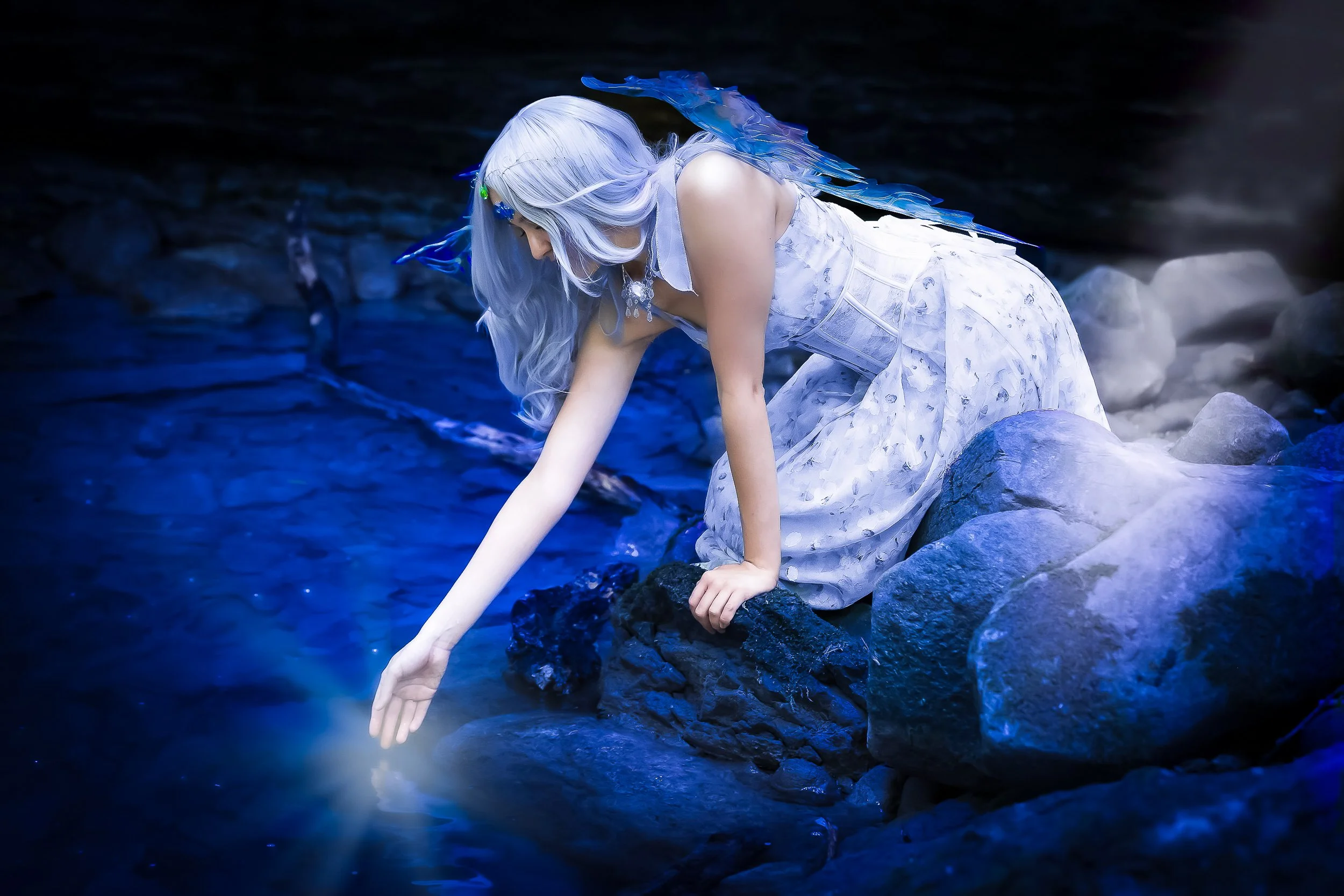 Une femme avec des cheveux argentés et des ailes bleues, vêtue d'une robe blanche, agenouillée sur des rochers près d'une rivière, touchant l'eau avec sa main dans une scène féerique.