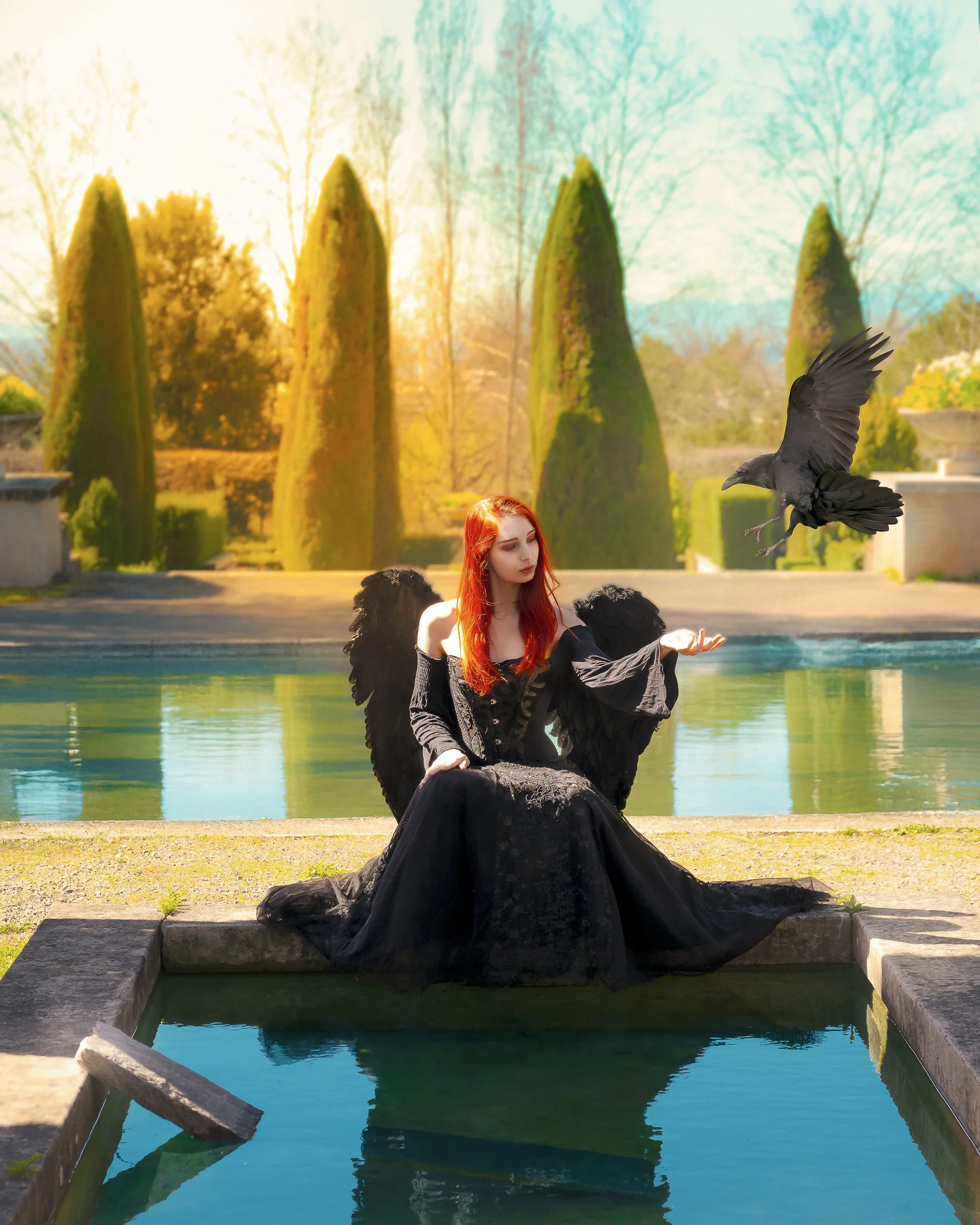 Une femme avec des ailes noires, habillée en noir, assise près d'une piscine, levant la main où un corbeau vole à proximité dans un jardin avec des arbres et des buissons verts.