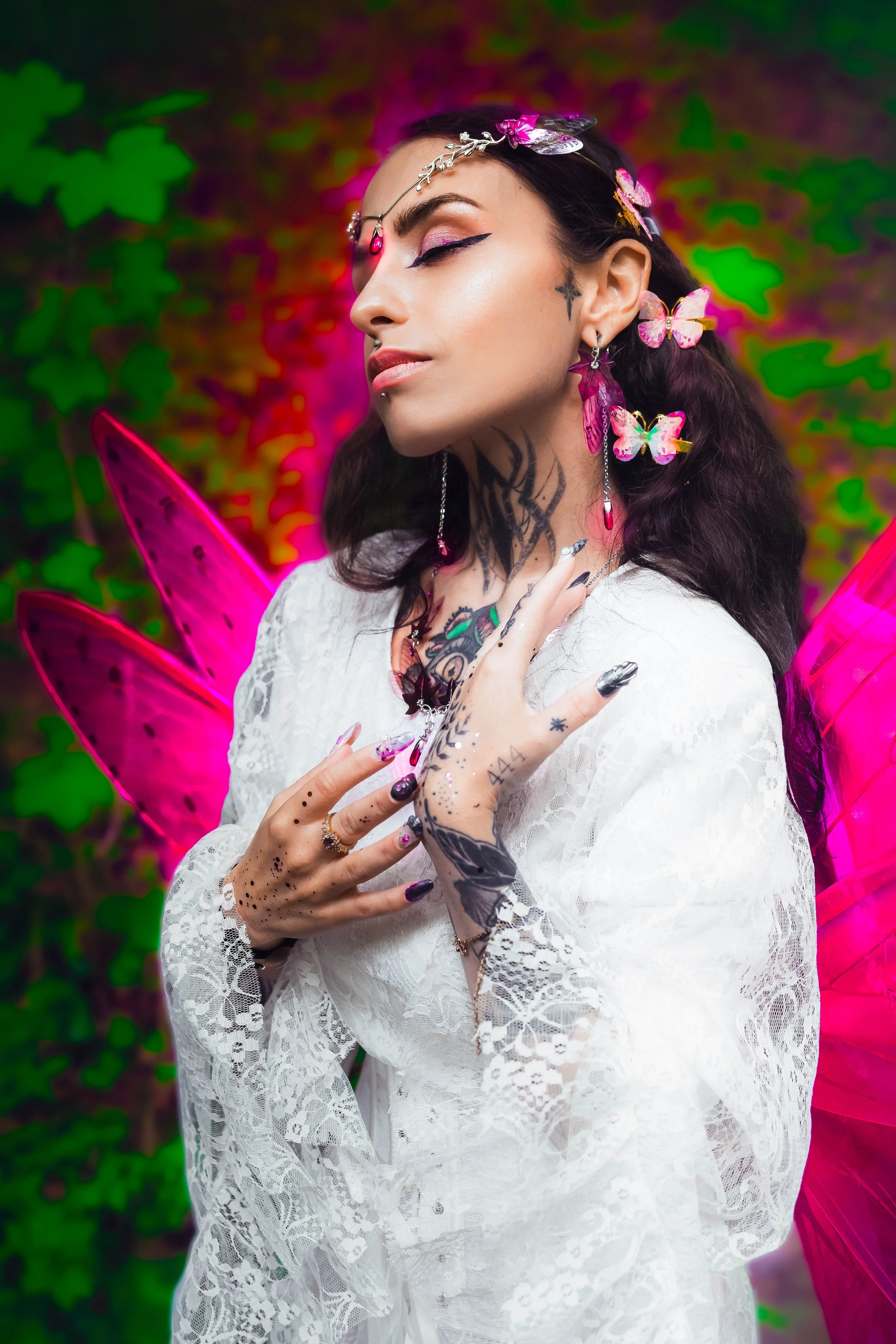 Une femme déguisée en fée avec des ailes roses, parée de bijoux et de tatouages, portant une robe blanche en dentelle, dans un décor coloré avec des feuilles vertes en arrière-plan.