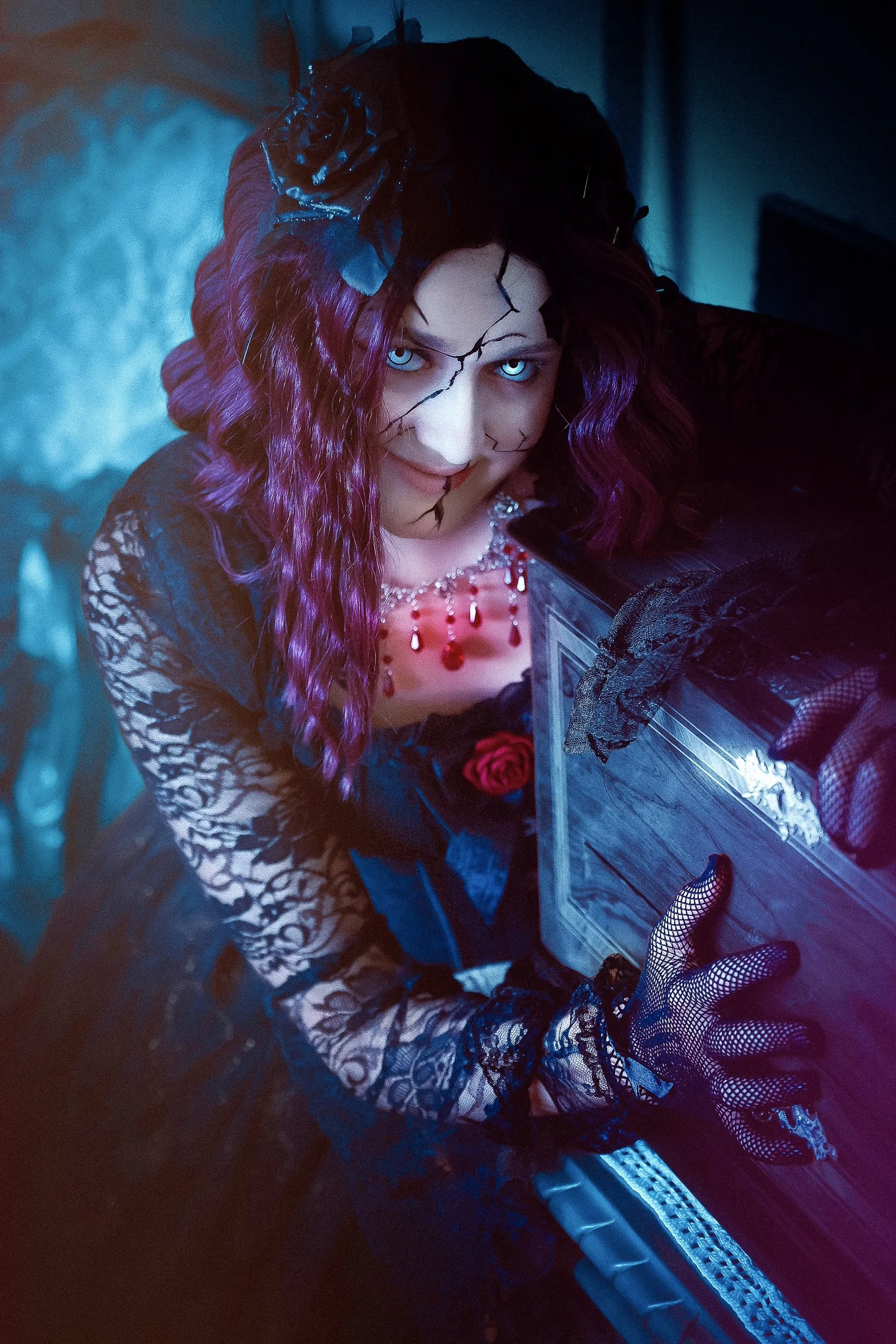 Une femme avec des cheveux violets et des fissures noires sur le visage porte une robe en dentelle noire, un collier de perles rouges, et des gants en filet noir, elle regarde vers le haut en souriant, dans une ambiance sombre et mystérieuse.