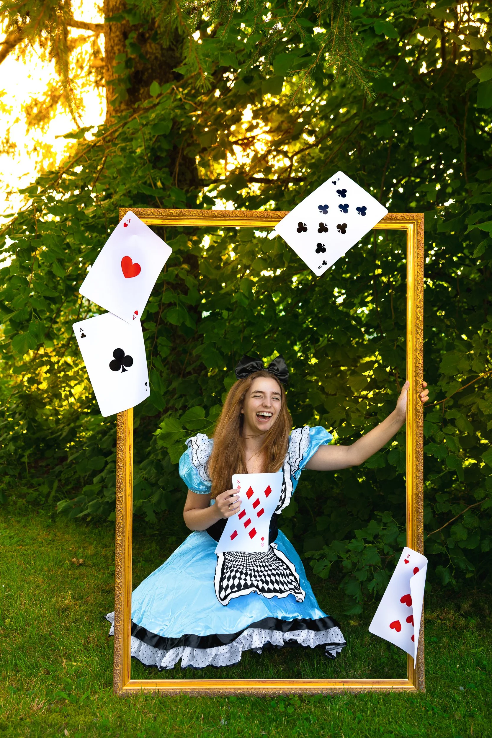 Une jeune femme habillée en Alice au pays des merveilles, souriante, tenant une carte de jeu, entourée de cartes flottantes, dans un cadre doré en plein air avec feuillage au soleil.